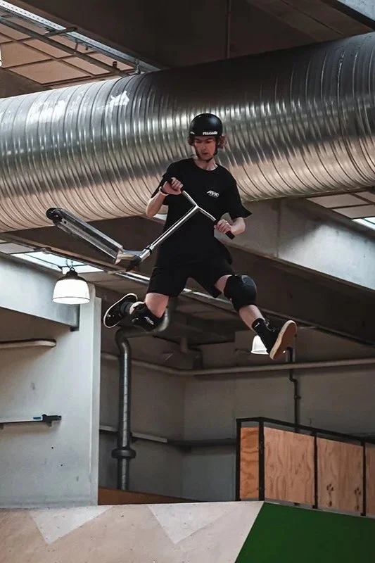 A person in protective gear, including helmet and knee pads, is mid-air performing a trick on a scooter in an indoor skate park.