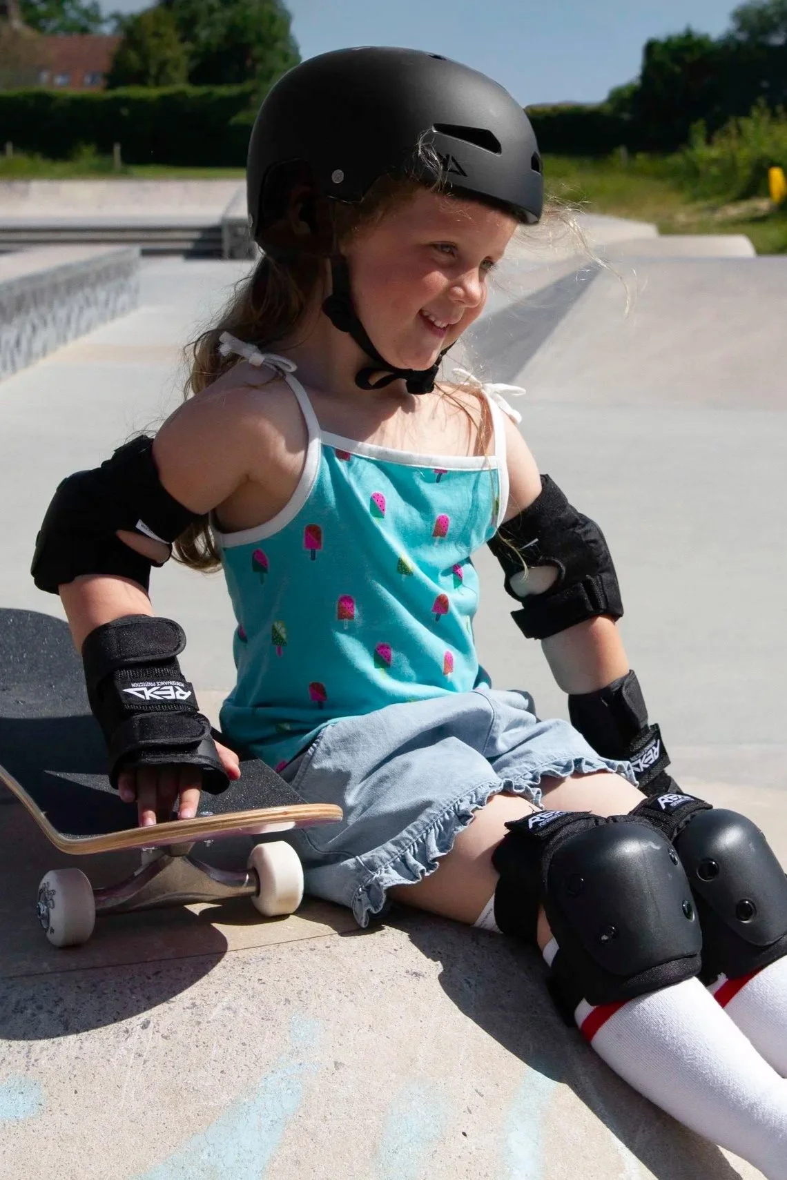 A young girl sitting on a skateboard at a skate park, wearing REKD protective gear including a helmet, elbow pads, knee pads, and wrist guards, smiling happily.