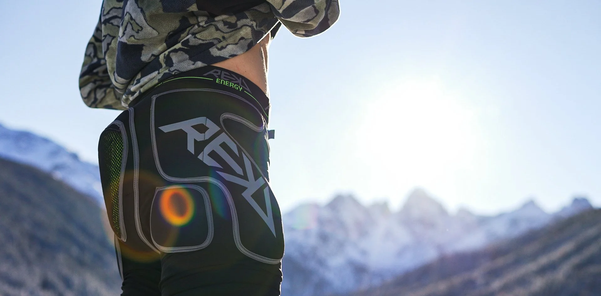 Close-up of a person's lower back wearing black and green athletic shorts with a geometric logo, standing outdoors in a snowy mountainous landscape with bright sunlight.