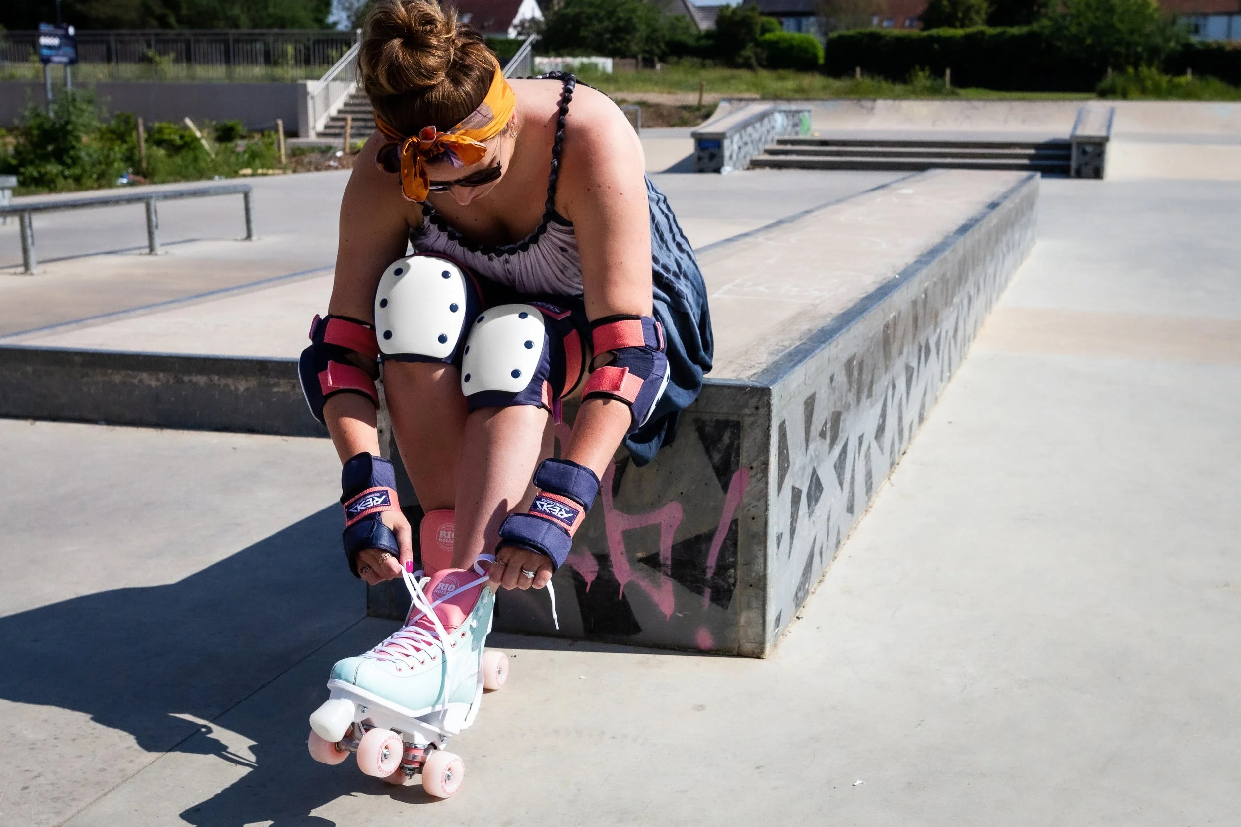 A woman with roller skates tying her shoelace at a skate park. She's wearing protective pads and a headscarf, with a smile on her face.