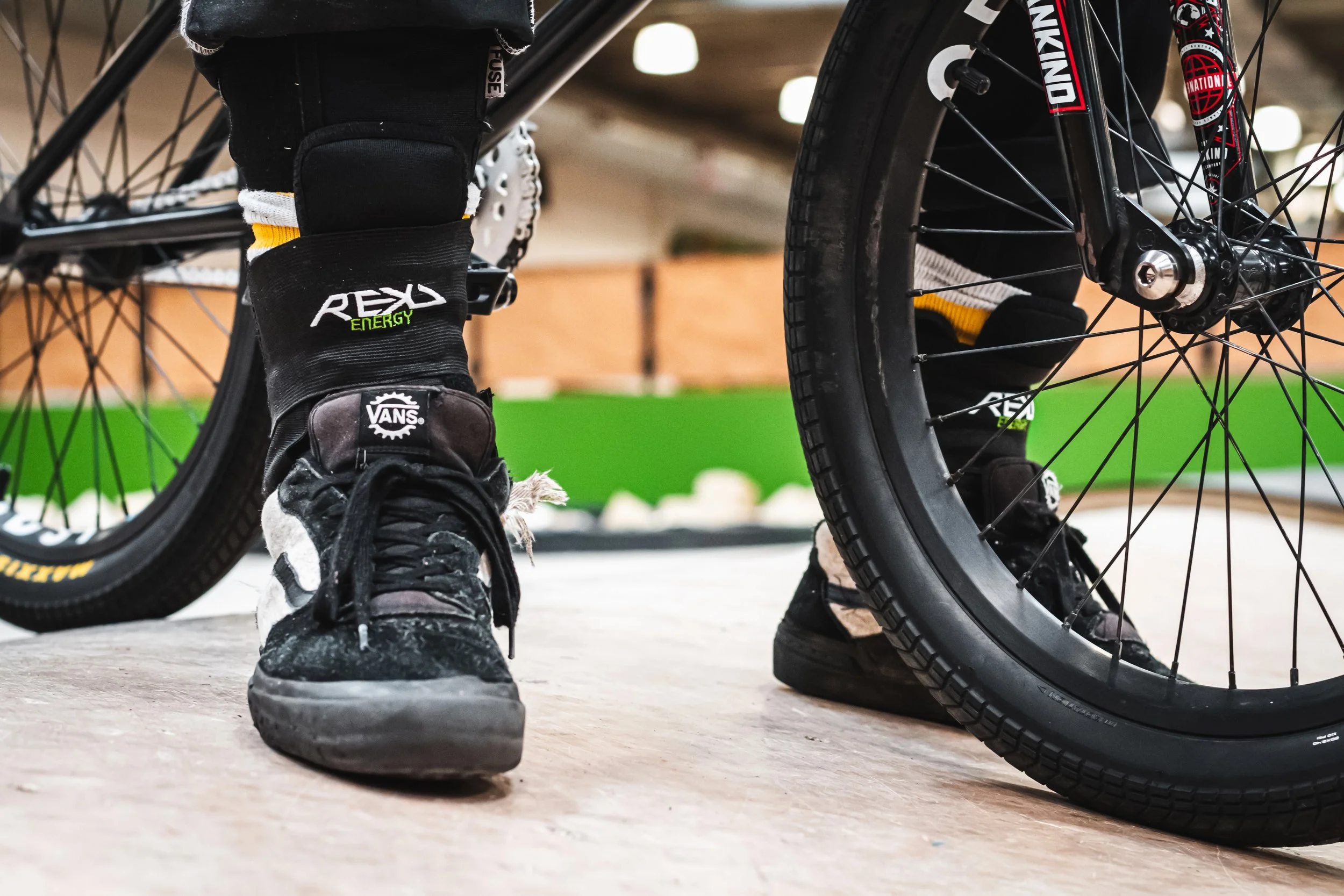 Close-up of a person wearing black sneakers with white and yellow socks, standing next to a mountain bike in an indoor setting.