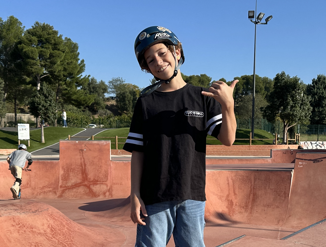 A boy wearing a helmet at a skate park, making a shaka sign with his right hand and smiling.