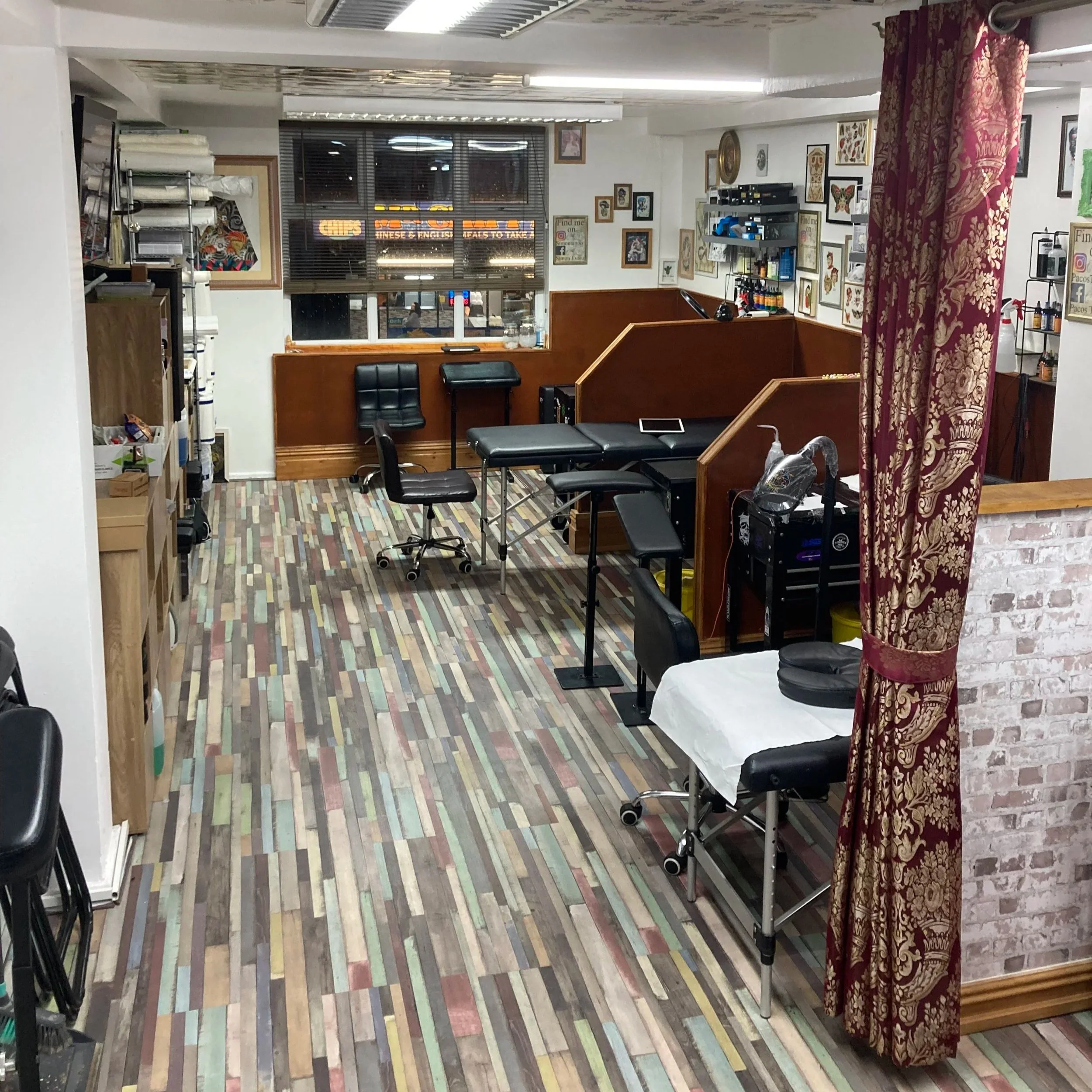 Interior of a tattoo shop with various artwork on the walls, chairs, shelves with supplies, a window with blinds, and a sari curtain on the right side.