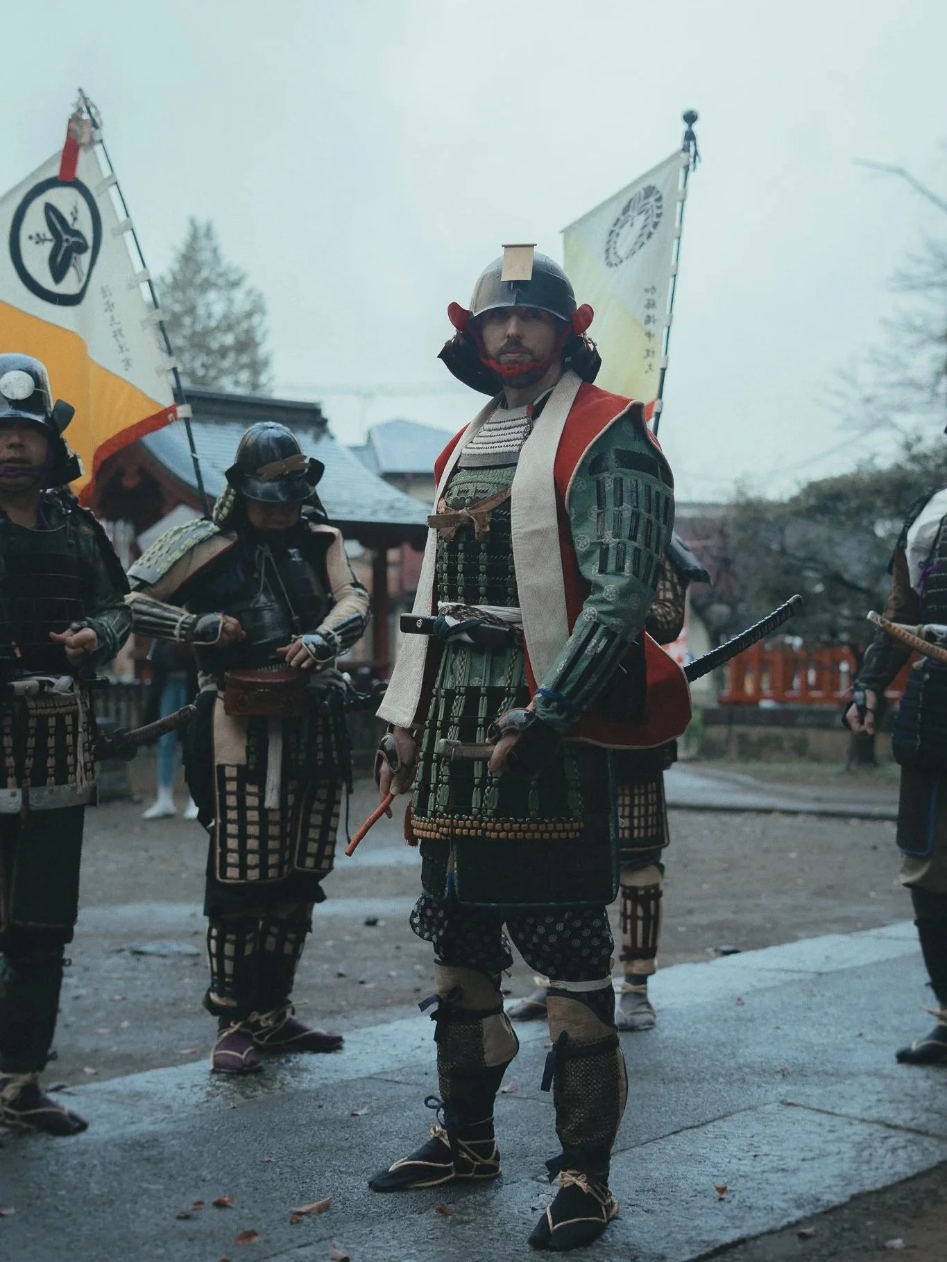Group of people dressed as samurai warriors holding flags, standing outdoors on a cloudy day, with traditional Japanese buildings in the background.