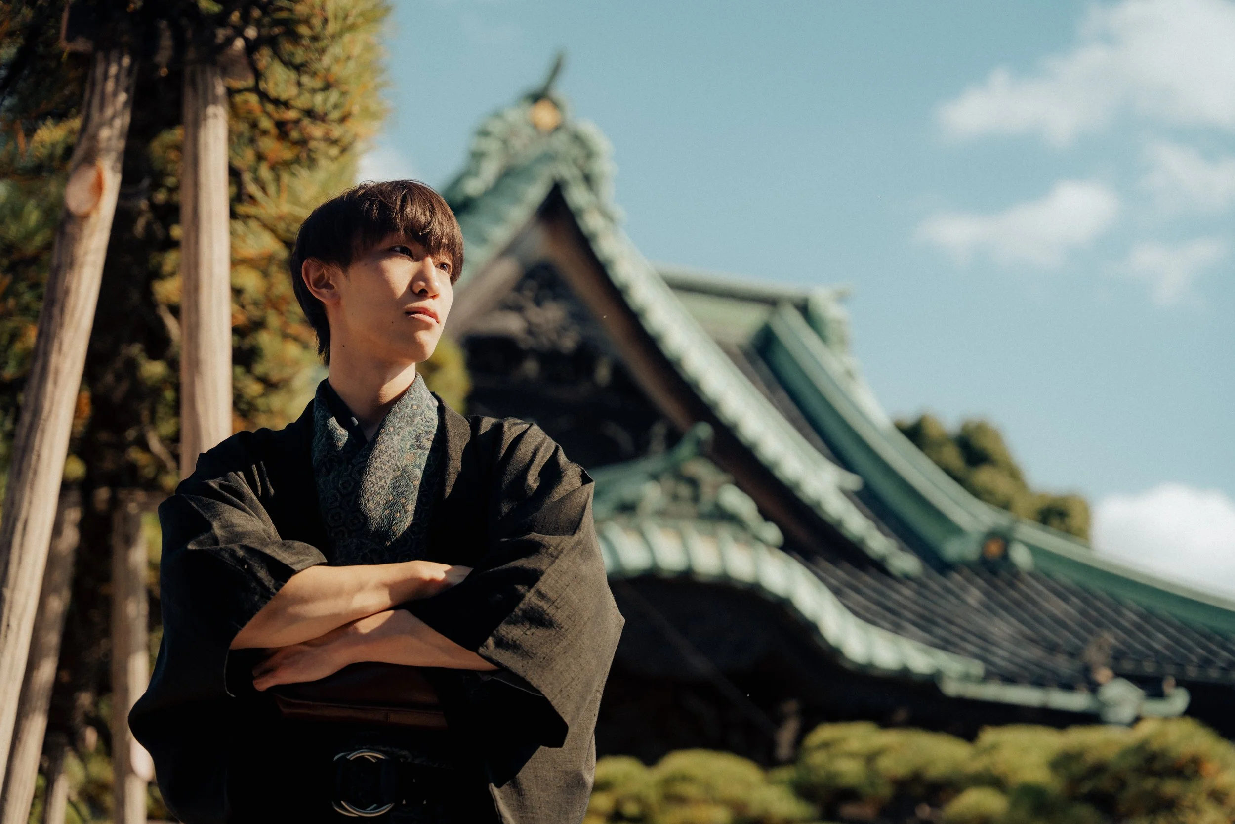 A young man in traditional Japanese attire standing outdoors with arms crossed, in front of a Japanese temple with a green tiled roof and wooden beams under a partly cloudy sky.