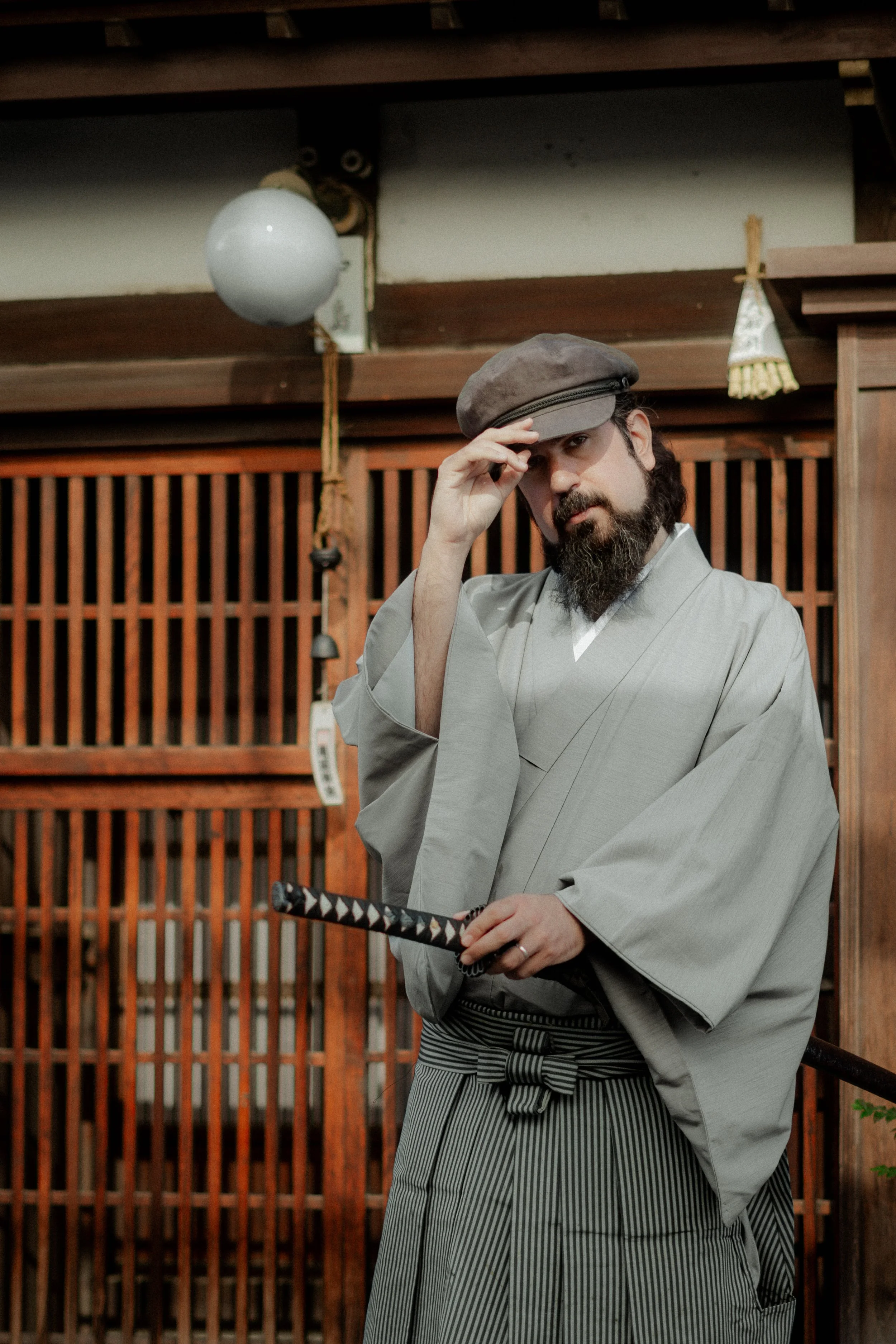 A man dressed in traditional Japanese attire, holding a katana, standing indoors with wooden paneling and decorations in the background.