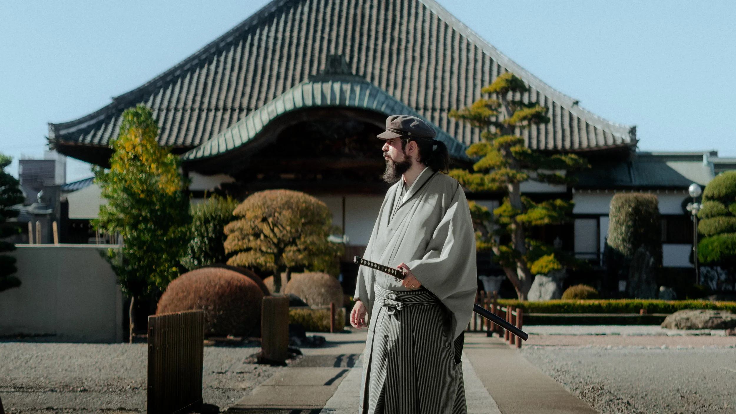 A man dressed in traditional Japanese attire, including a kimono and a hat, holding a katana sword, standing in front of a Japanese temple with a tiled rooftop and landscaped garden with trimmed bushes.