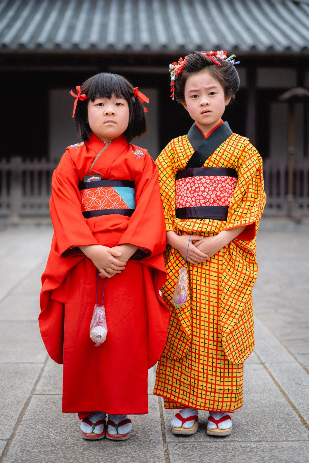 Two young girls wearing traditional Japanese kimono, standing outside in front of a building with a tiled roof, both with serious expressions and wearing zori sandals.
