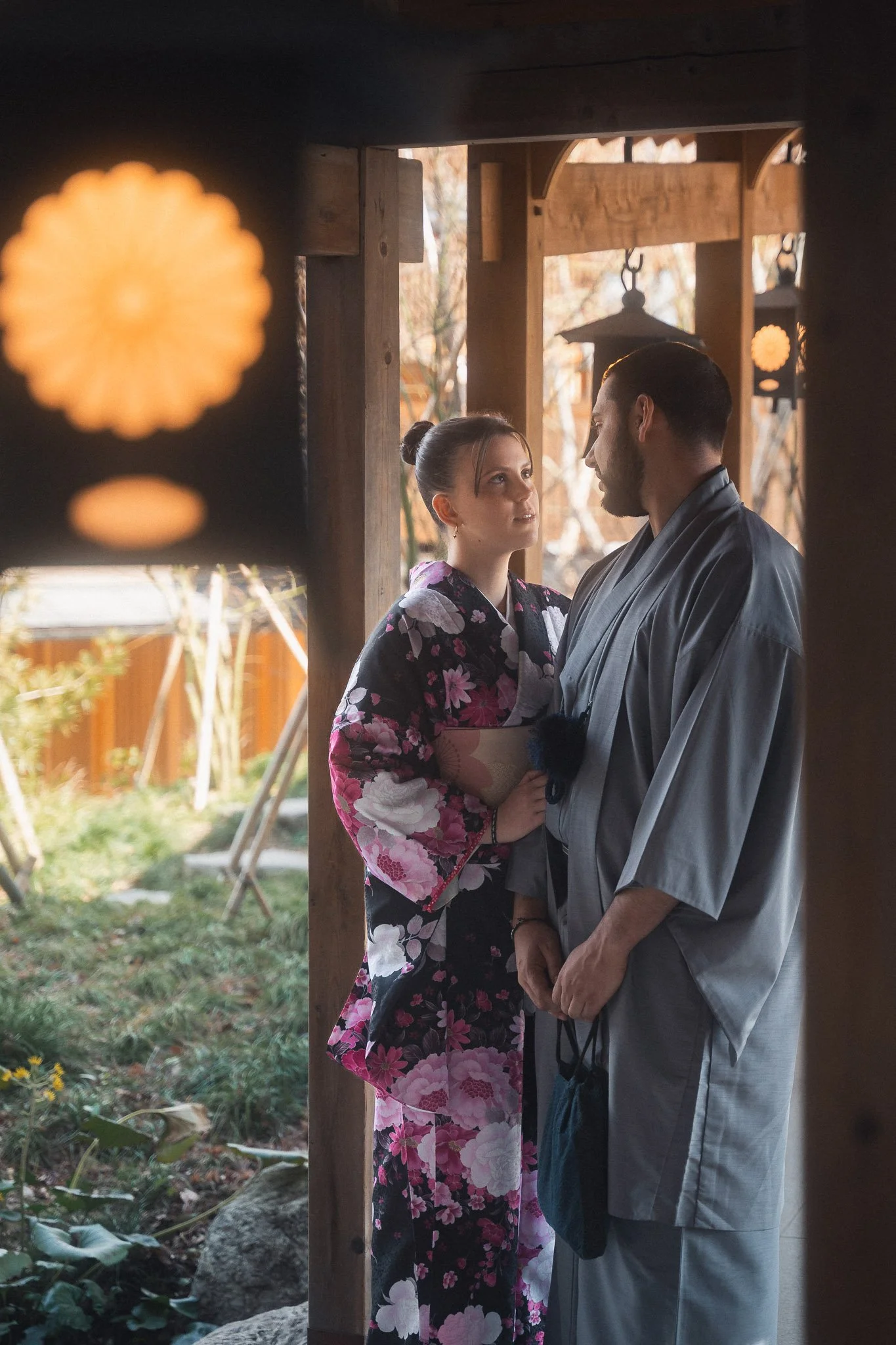 A woman and a man face each other closely, standing under a wooden structure, both dressed in traditional Japanese attire. The woman is wearing a colorful kimono with pink, purple, and white flowers, and the man is dressed in gray robes. They are looking into each other's eyes, with outdoor trees and wooden elements visible in the background.