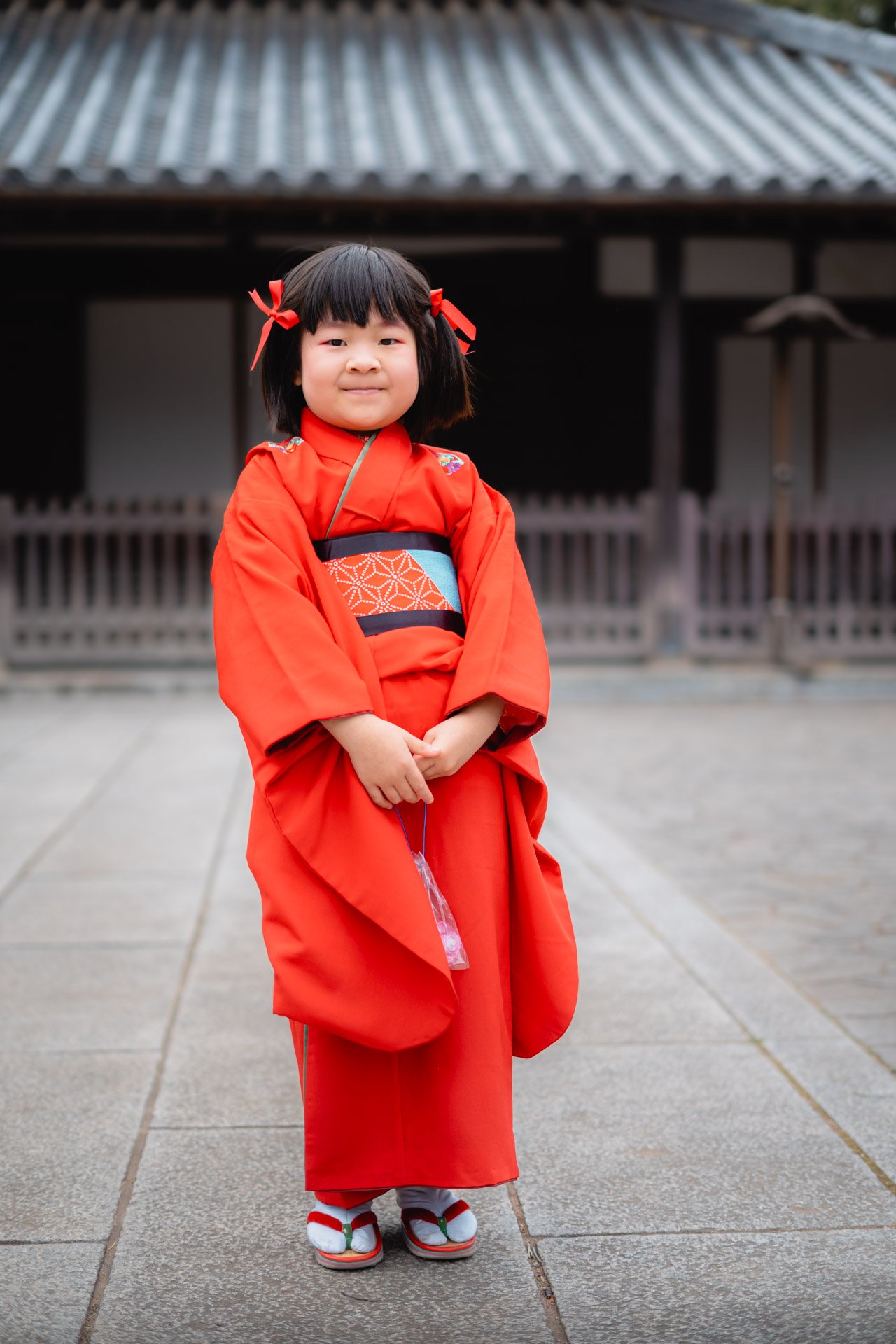 Young girl wearing a red kimono with traditional Japanese sandals, standing outdoors in front of a building with a wooden fence and tiled roof.