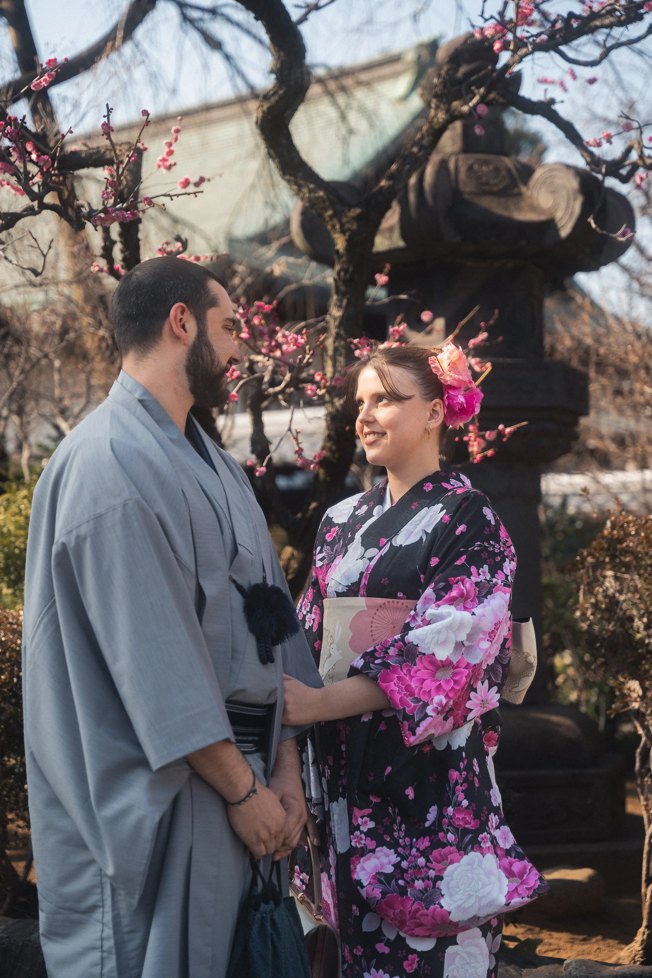 A man and a woman dressed in traditional Japanese kimonos are standing outdoors under a blooming cherry blossom tree, gazing at each other.