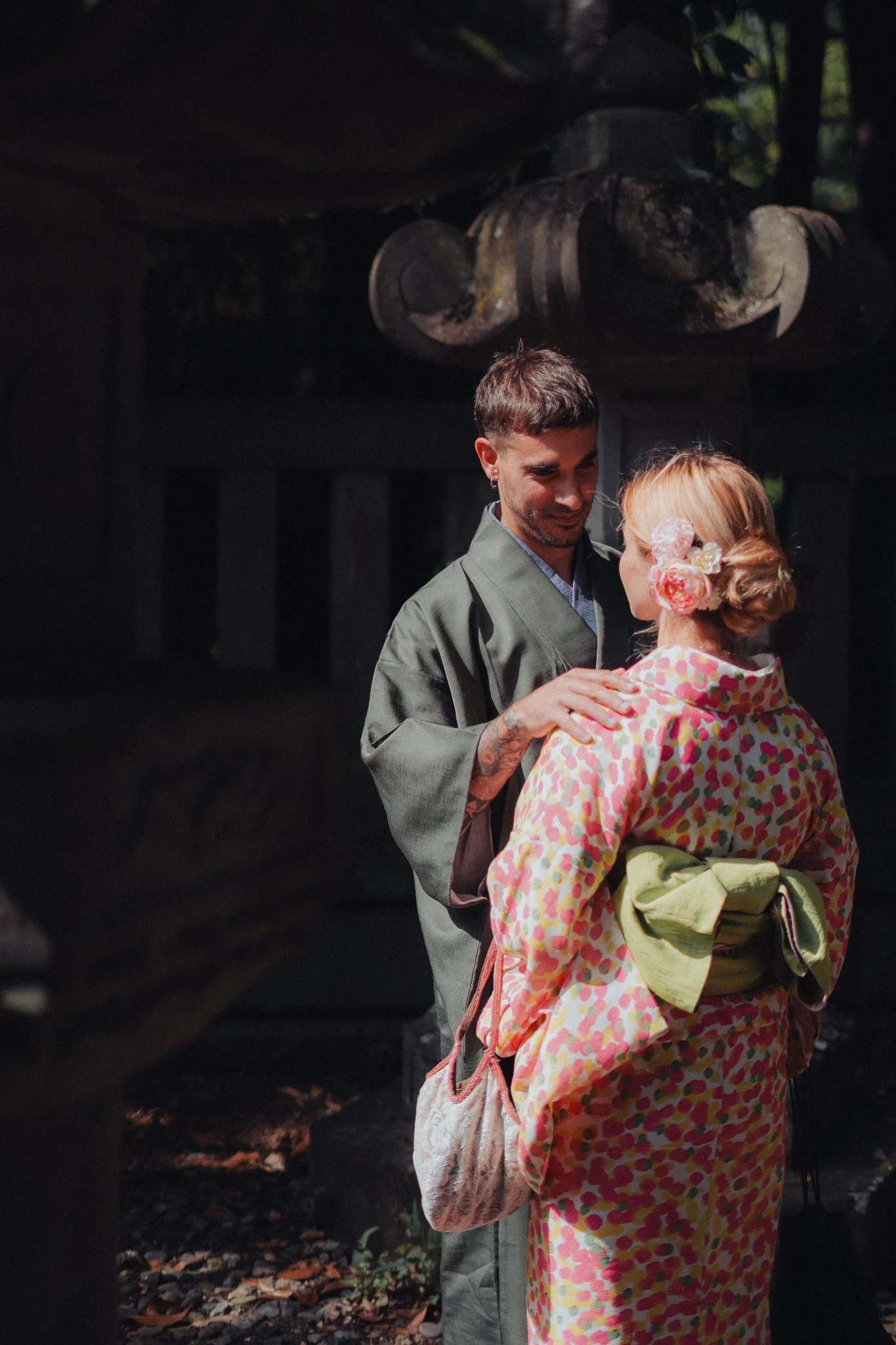 A man and woman stand close to each other, with the man gently holding the woman's shoulder, outdoors in a shaded area with fallen leaves on the ground, and dark wooden structure in the background.
