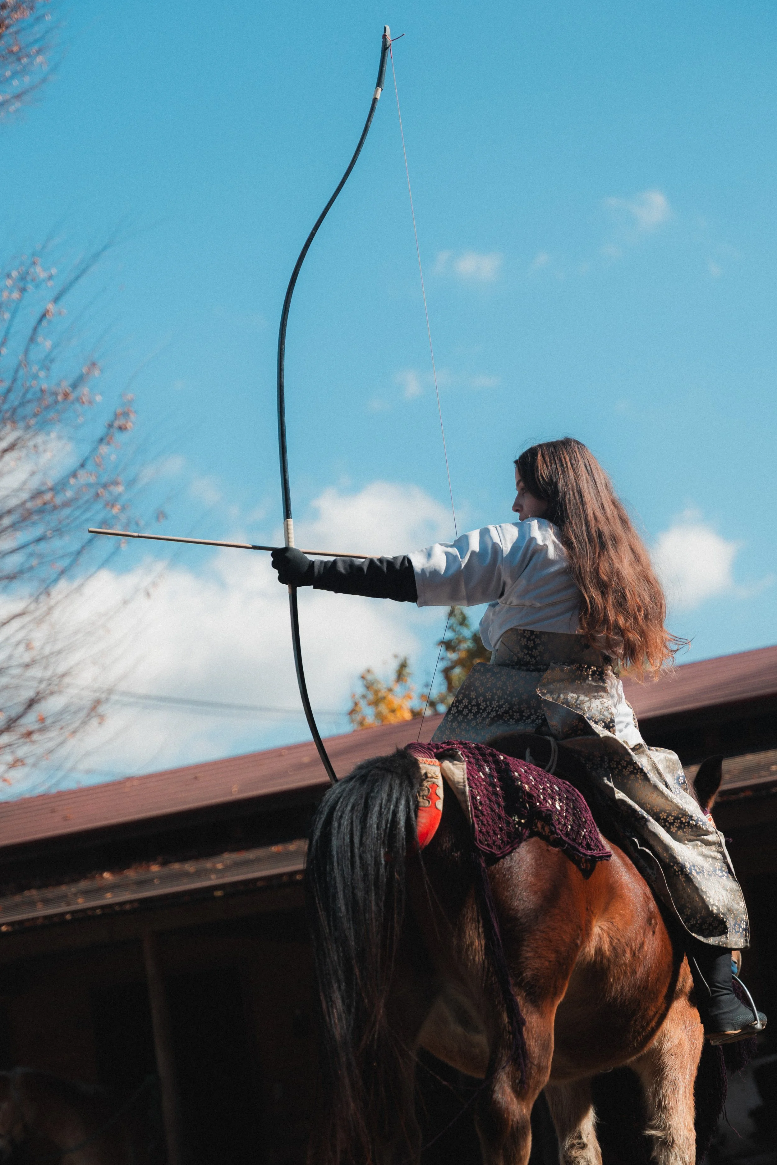 A woman in traditional clothing is riding a horse and aiming a bow and arrow at the sky, with a clear blue sky and few clouds in the background.