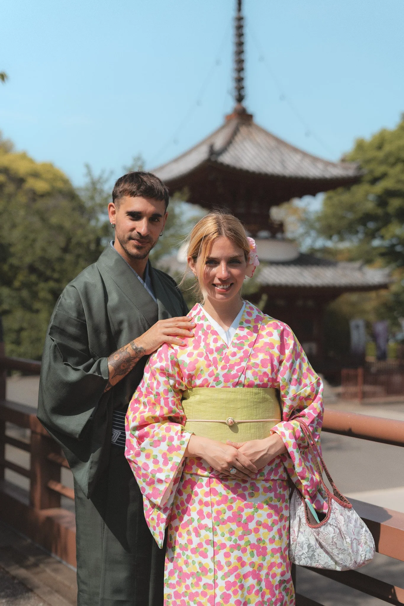 A man and a woman stand outdoors with a traditional Japanese pagoda in the background on a sunny day. The man has short dark hair, tattoos on his hand, and is dressed in a gray kimono. The woman has blonde hair with a flower accessory, and is wearing a colorful kimono with pink, green, and yellow patterns, with her hands clasped in front of her.