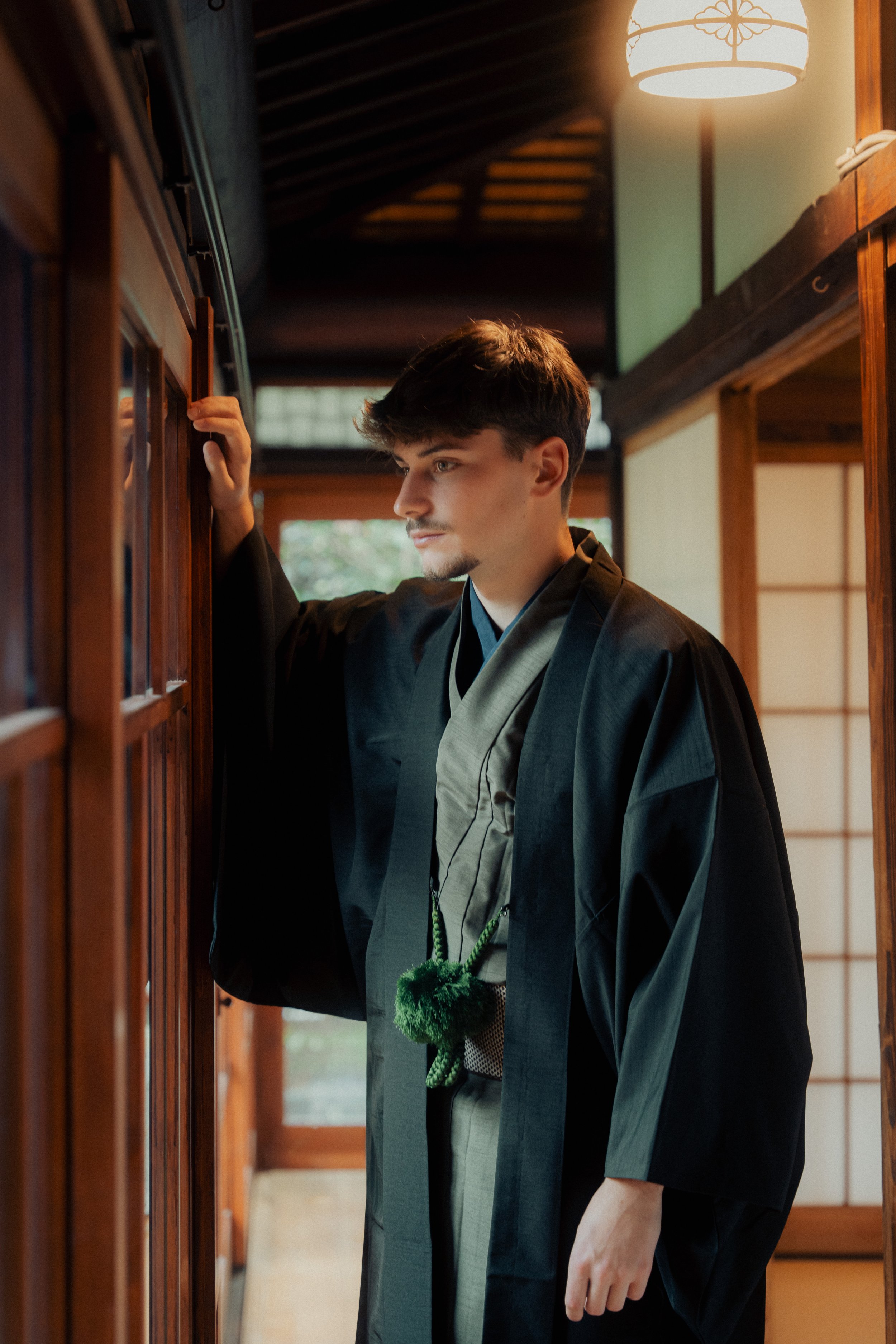 A man dressed in traditional Japanese attire, standing in a wooden interior with sliding shoji doors and a paper lantern hanging from the ceiling. The man is looking at a wooden display case with glass panels, possibly in a museum or cultural center.