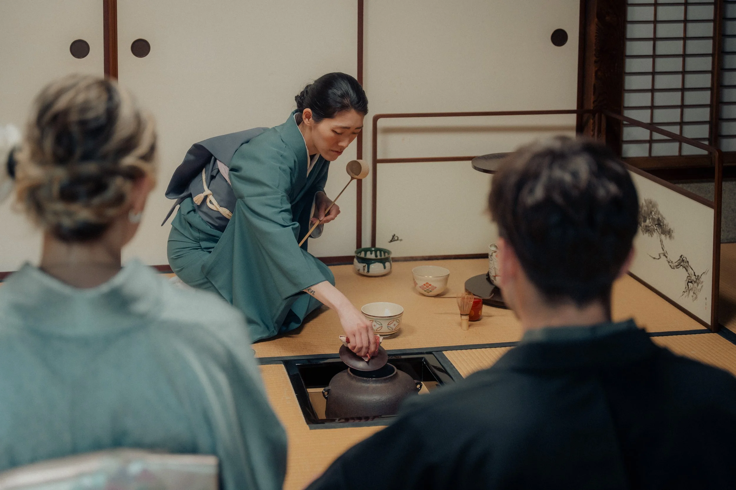 A woman in traditional Japanese attire demonstrates tea ceremony to people seated around a low table in a Japanese-style room.