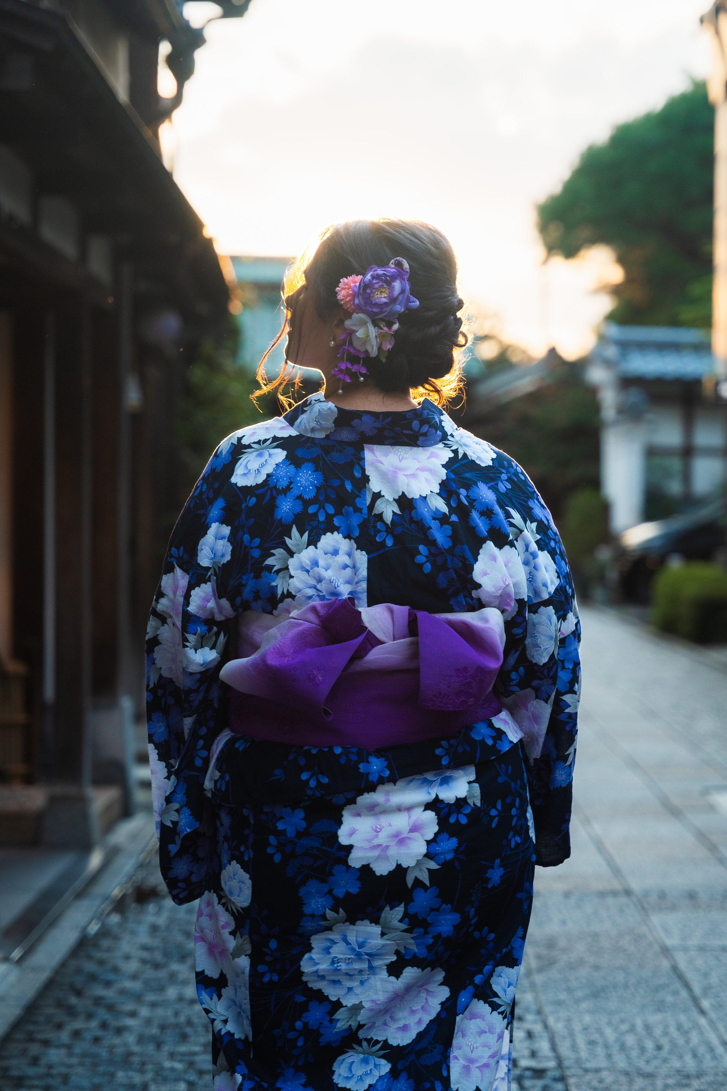 A woman wearing a traditional Japanese yukata with blue and white floral patterns, with a purple obi, and adorned with purple and pink flowers in her hair, walking outdoors during sunset.