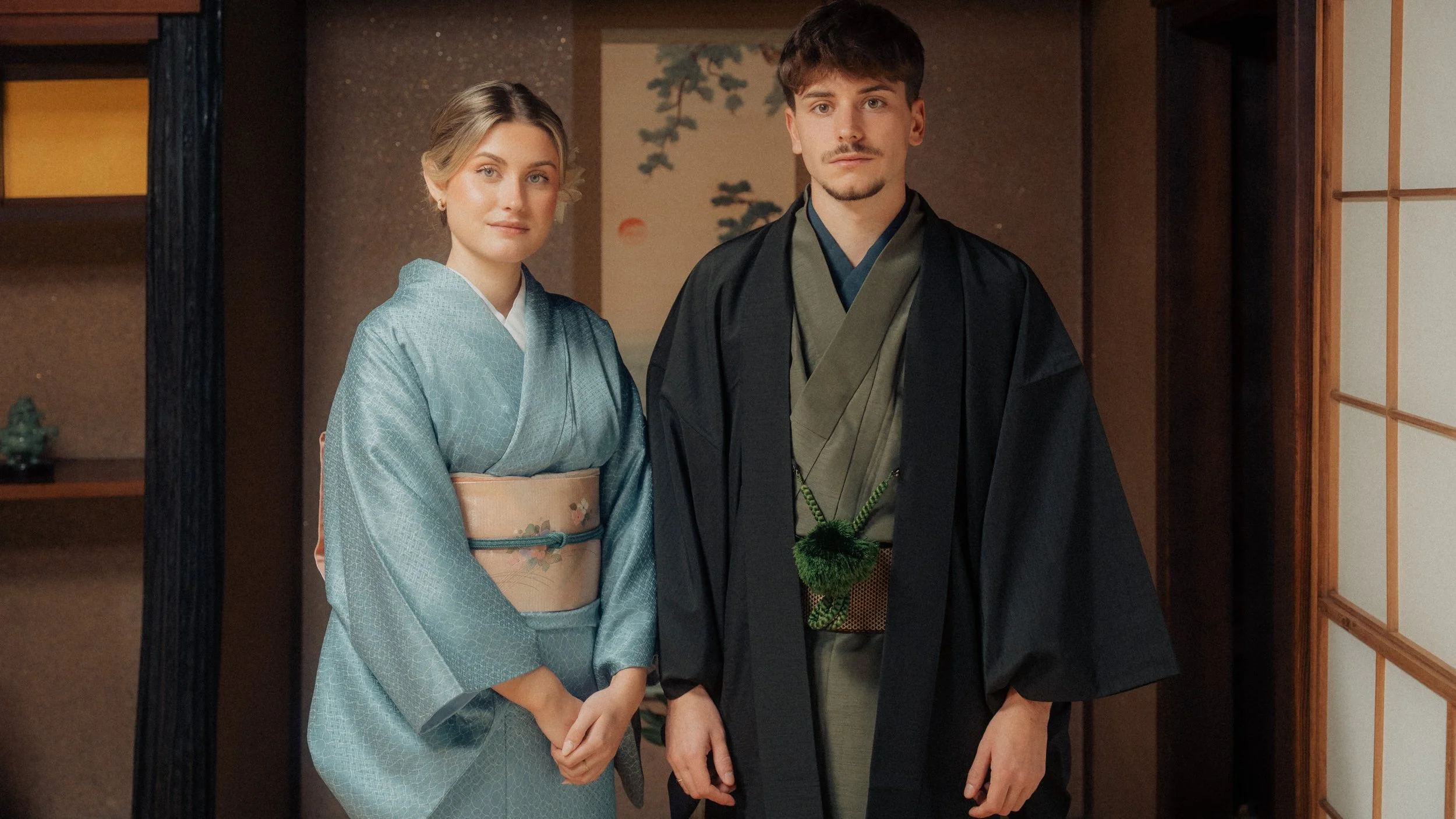 A woman and a man dressed in traditional Japanese kimonos standing inside a room with kanji artwork and shoji doors in the background.