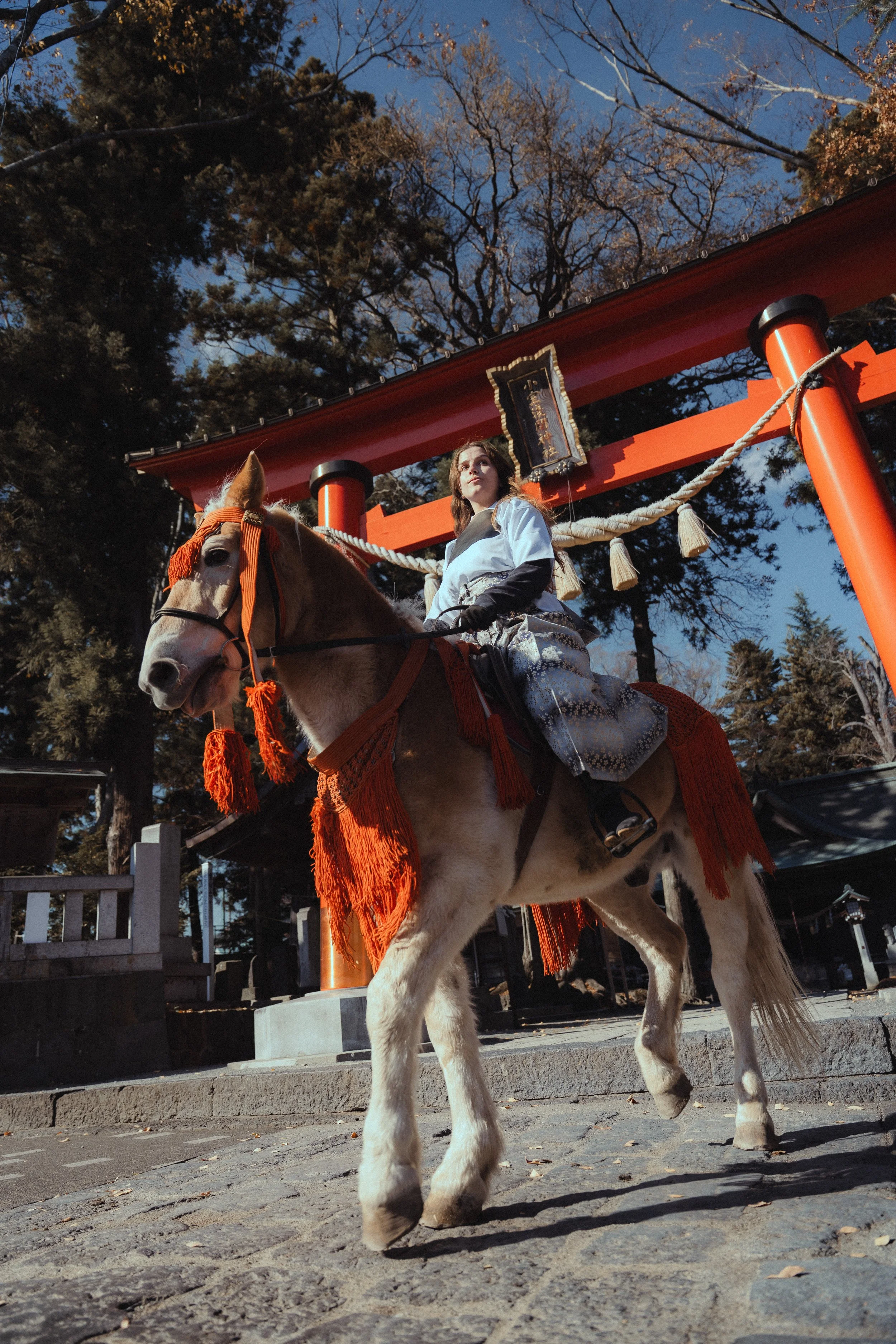 A woman dressed in traditional Japanese clothing riding a white horse decorated with orange tassels and harness. They are positioned in front of a large red Torii gate at a Shinto shrine, with tall trees and a clear blue sky in the background.