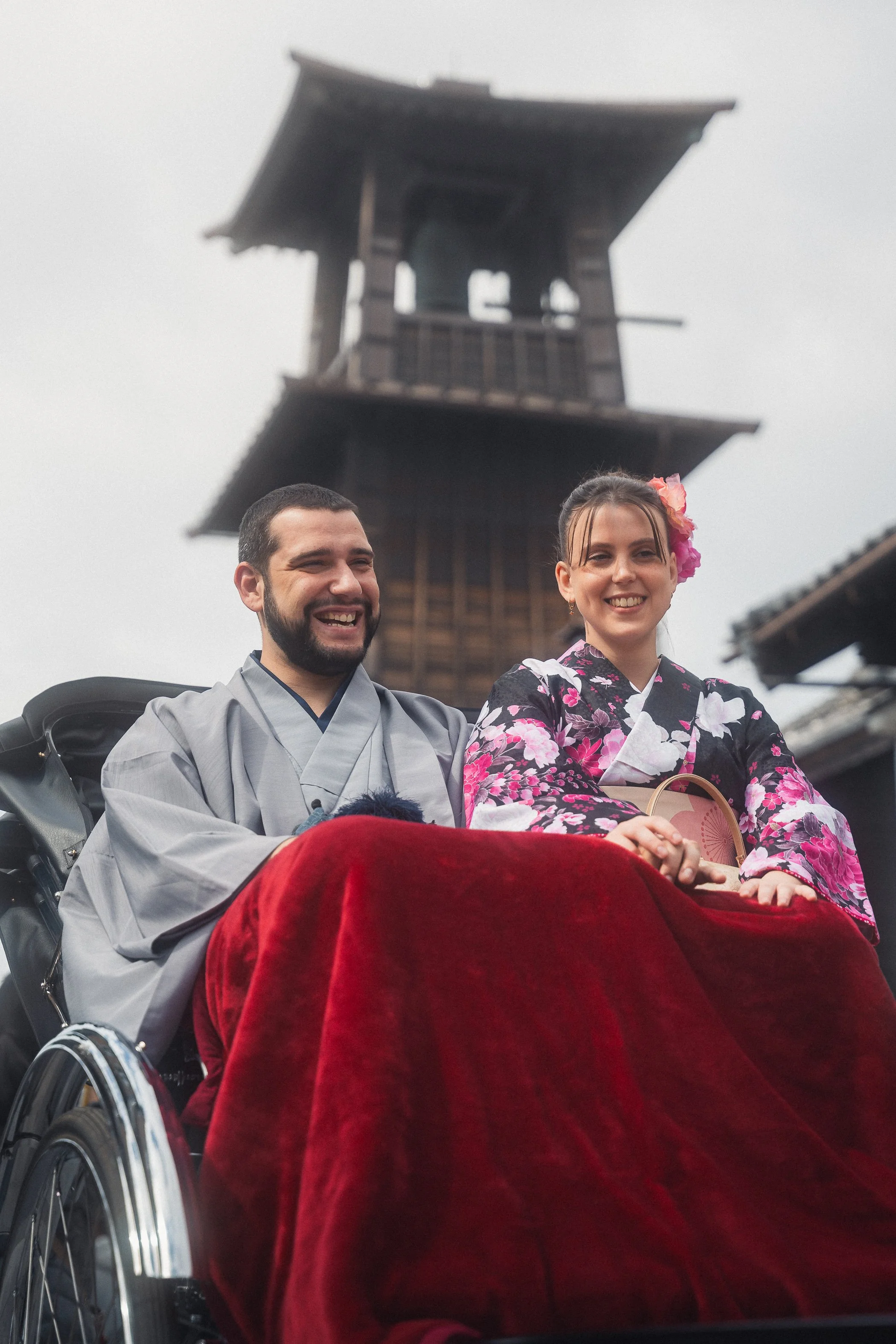A smiling man and woman in traditional Japanese attire sitting together on a red velvet blanket in front of a wooden pagoda-style structure.