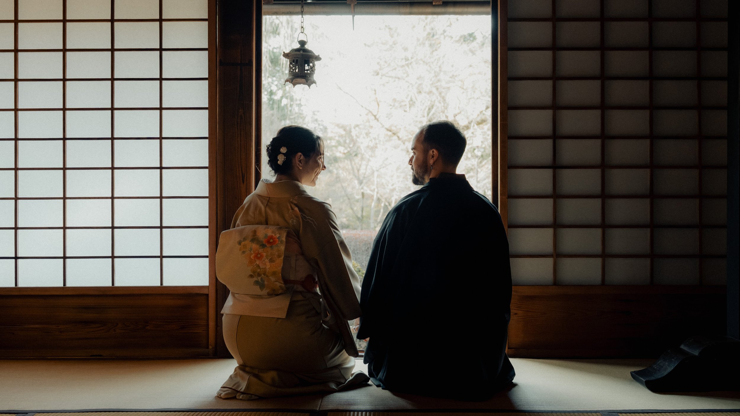 A couple dressed in traditional Japanese clothing sitting on tatami mats and facing each other inside a room with shoji screens and a view of trees outside.