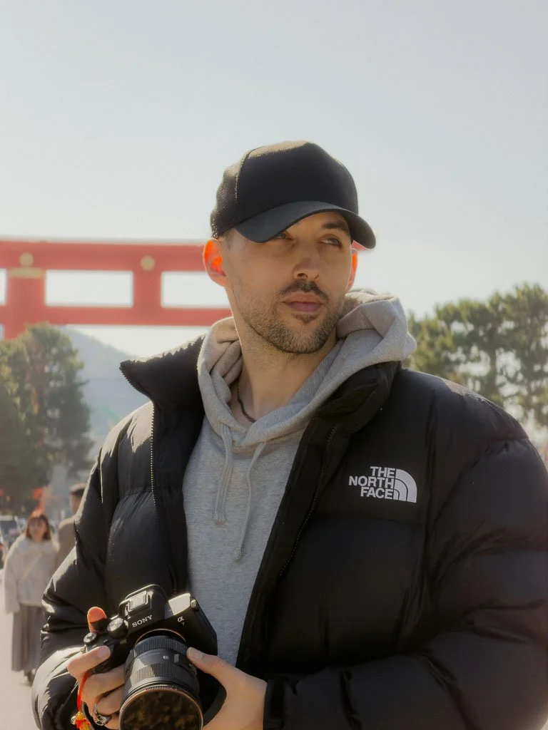 A man wearing a black cap, black puffy jacket, and gray hoodie holding a camera, outdoors near a red torii gate and trees.