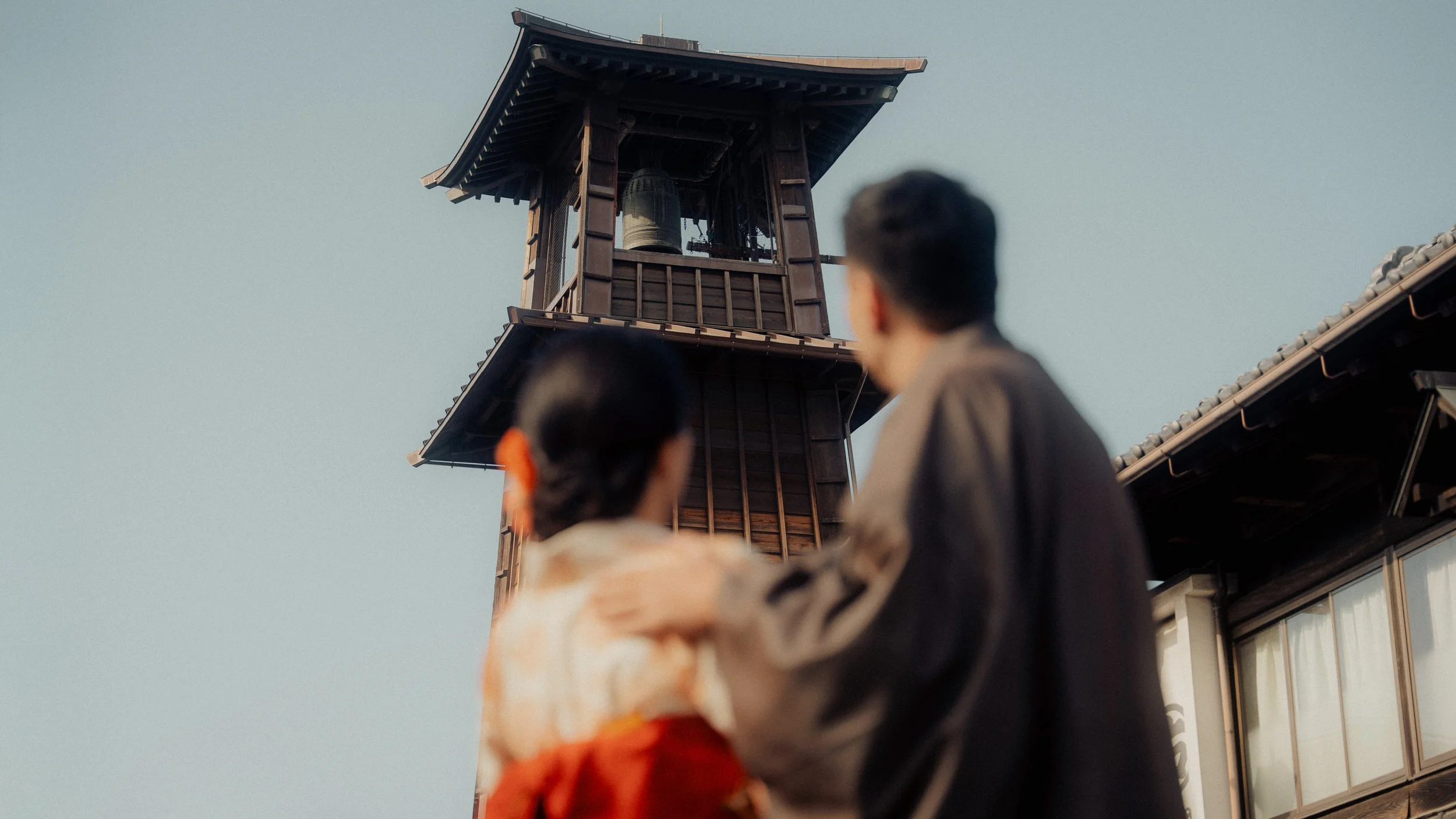 Two people, a man and a woman, are walking outdoors near a wooden clock tower with a bell. The scene appears to be in a residential area with traditional-style buildings.