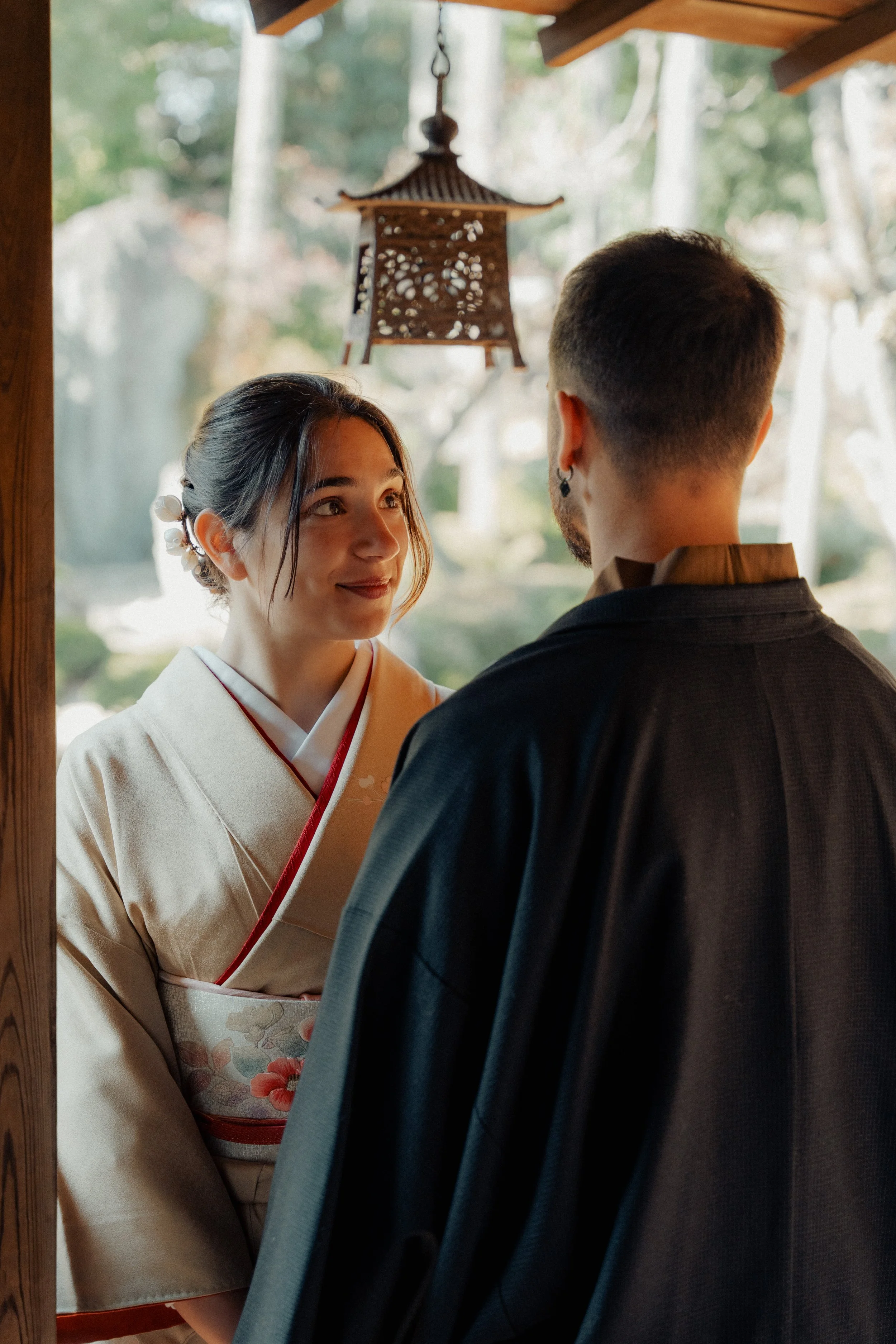 A woman dressed in a traditional Japanese kimono standing in front of a mirror, looking at a man with short dark hair and an earring, who is facing away from the camera. The background features blurred greenery and a hanging wooden lantern.