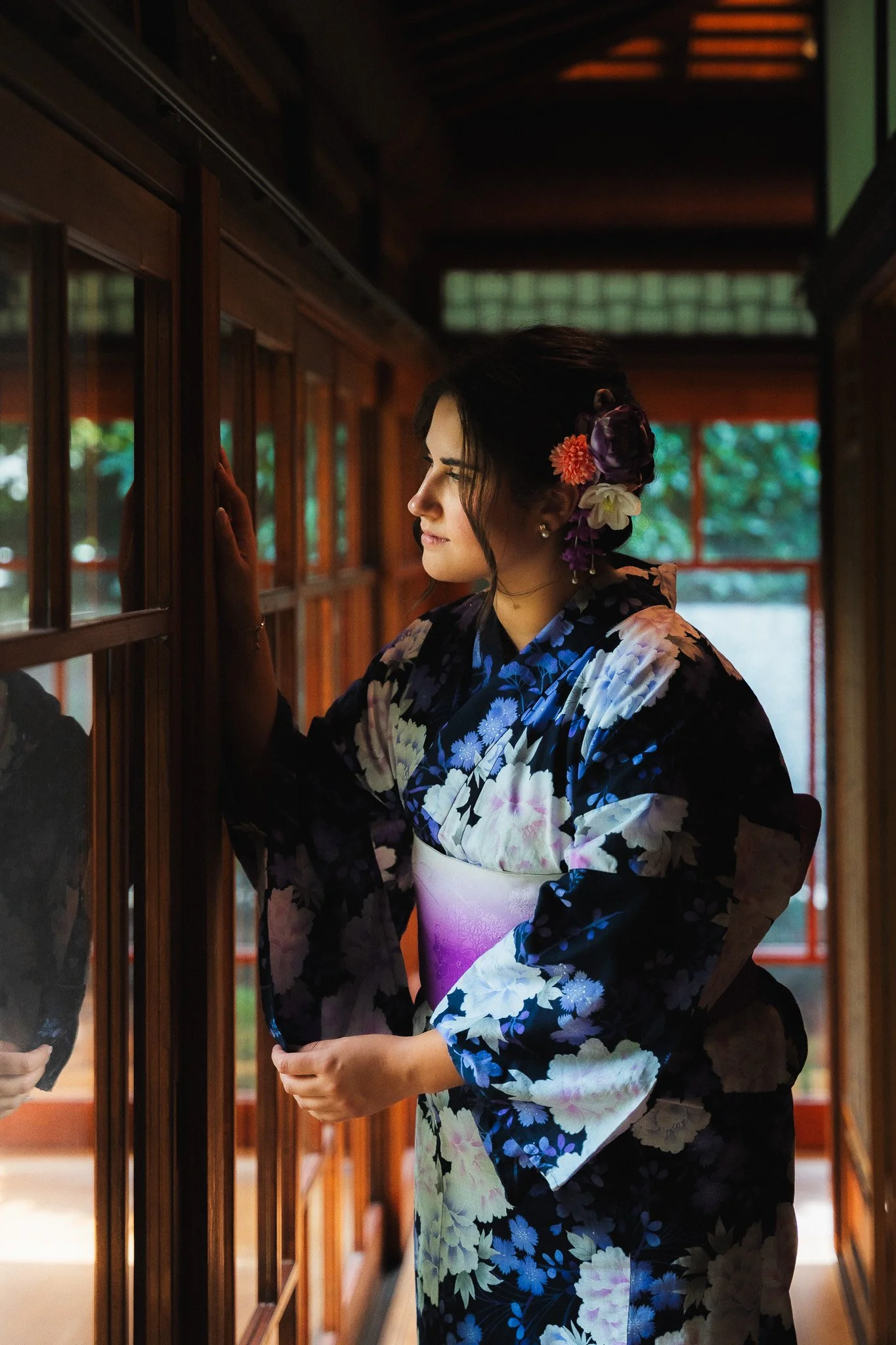 A woman dressed in a traditional Japanese kimono with floral patterns, adorned with colorful flowers in her hair, standing inside a wooden building with shoji screens and looking through a glass window.