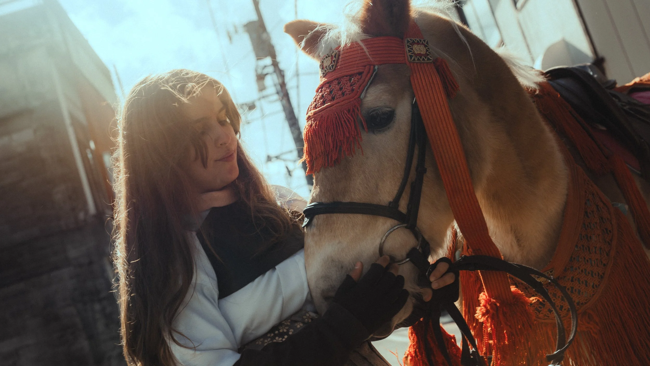 A girl with long brown hair and a white jacket helping a horse put on an orange decorative harness. The horse has a tan coat with a white mane and is wearing a colorful bridle with embroidery. The scene takes place outdoors, with sunlight and buildings in the background.