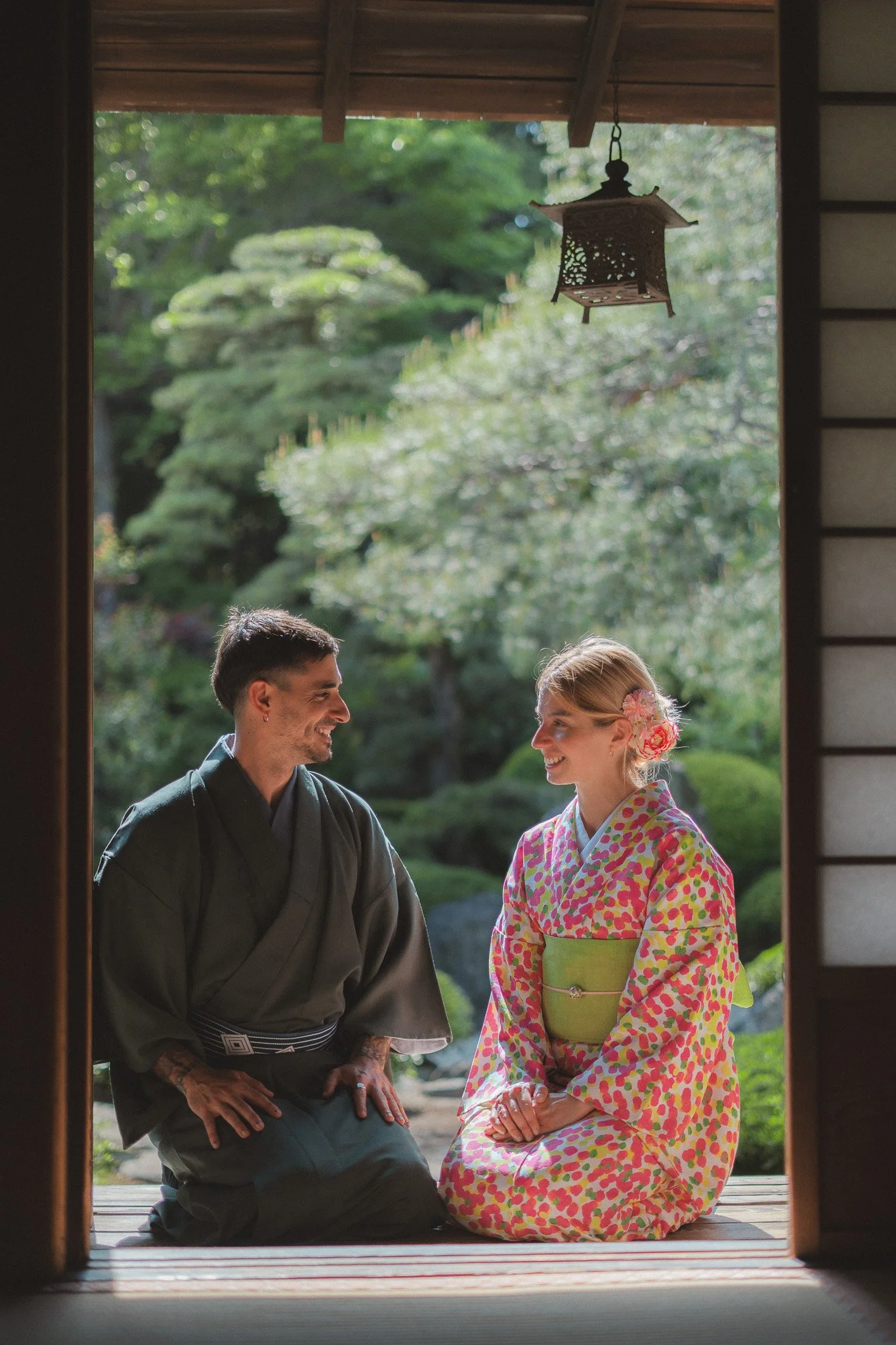 A man and a woman dressed in traditional Japanese kimonos, sitting on a wooden porch, smiling and looking at each other, with a lush garden and trees in the background, visible through the open doorway.
