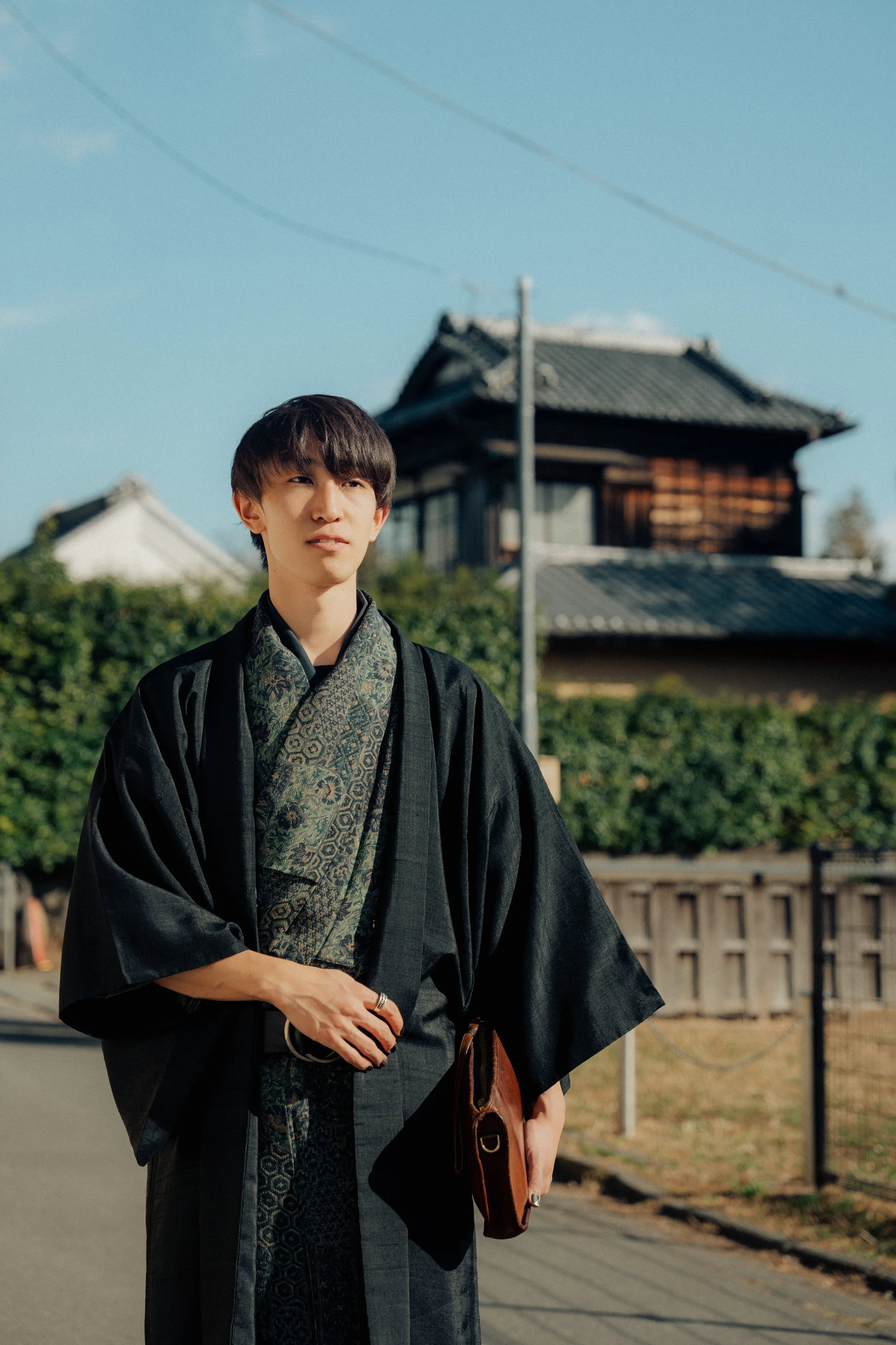 A young man wearing traditional Japanese clothing, standing outdoors near a traditional Japanese building with a tiled roof, under a clear blue sky.
