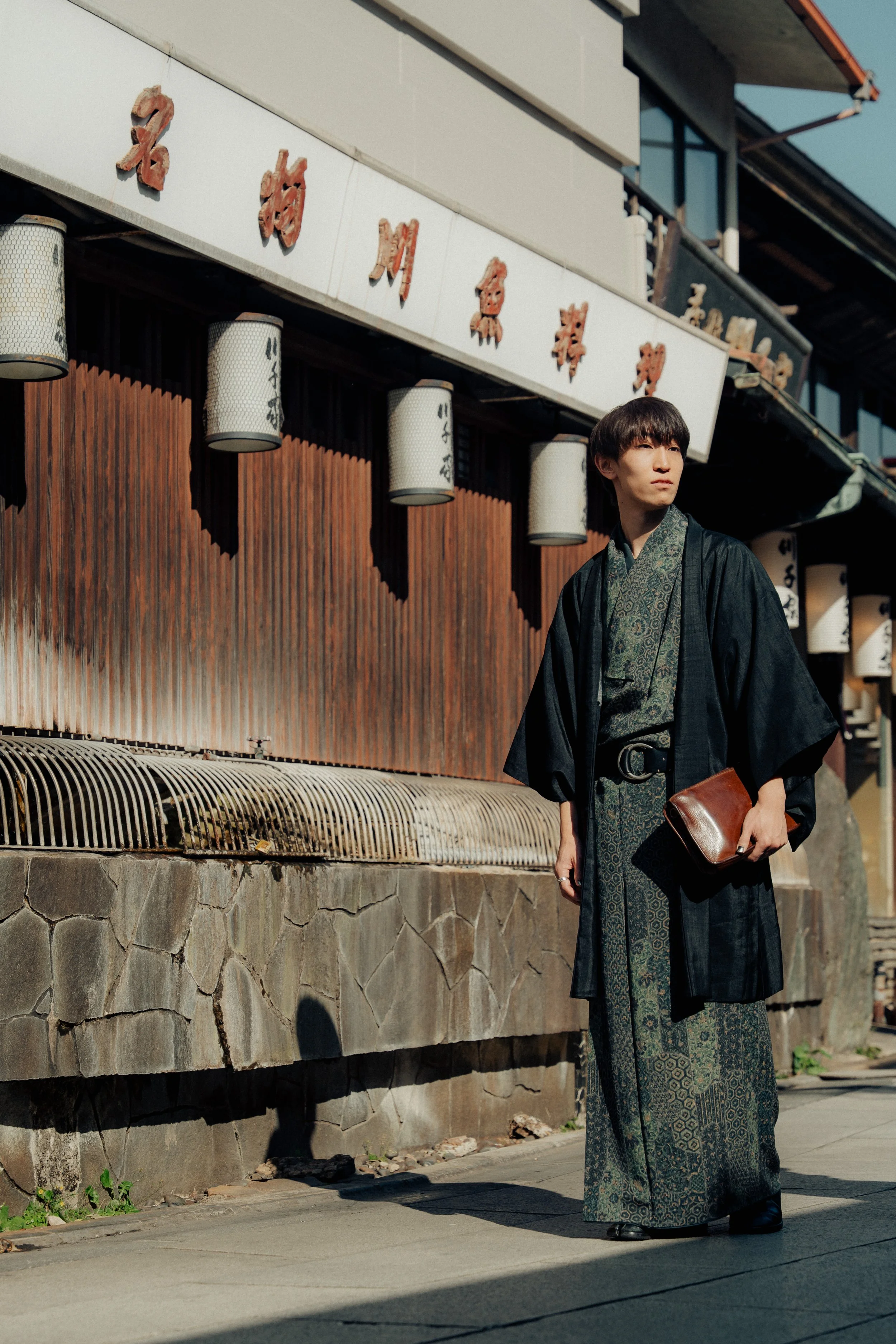 A young man dressed in traditional Japanese clothing, standing on a sidewalk in front of a wooden building with Japanese signs and lanterns, under clear blue skies.