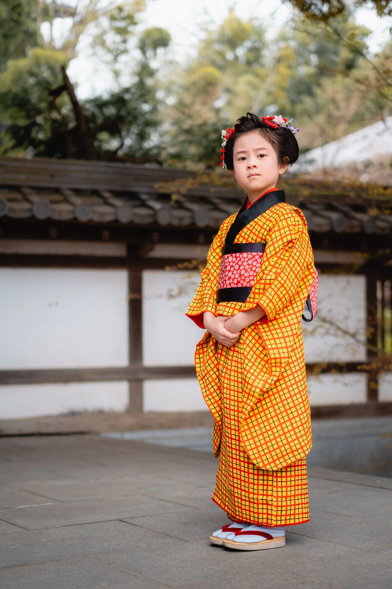 Young girl dressed in traditional Japanese kimono standing outdoors with a serene background of trees and a wooden fence.