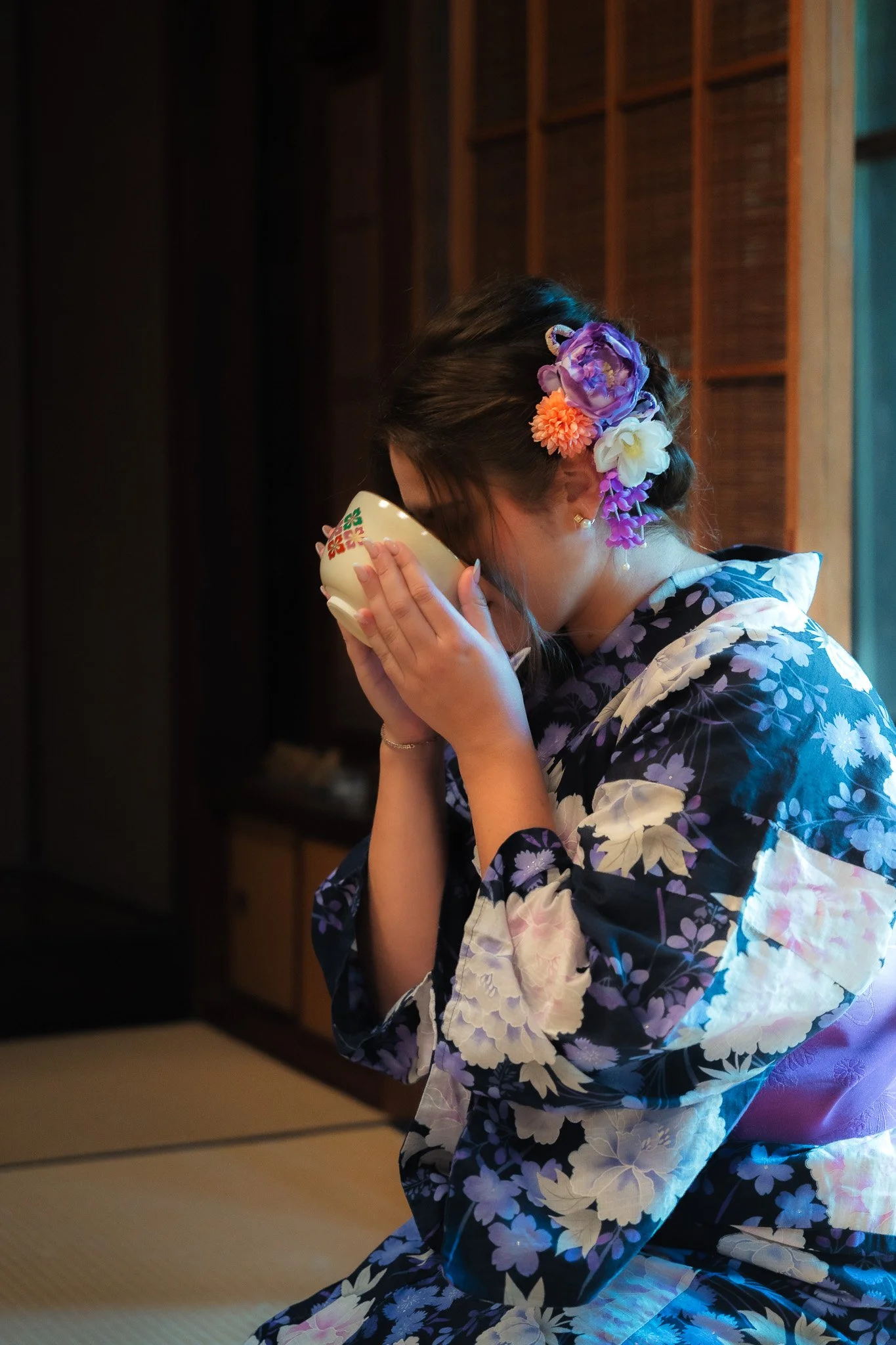 A woman dressed in a traditional Japanese kimono with floral patterns, kneeling on tatami mats, holding a tea cup close to her face during a tea ceremony, with her hair decorated with colorful flowers.