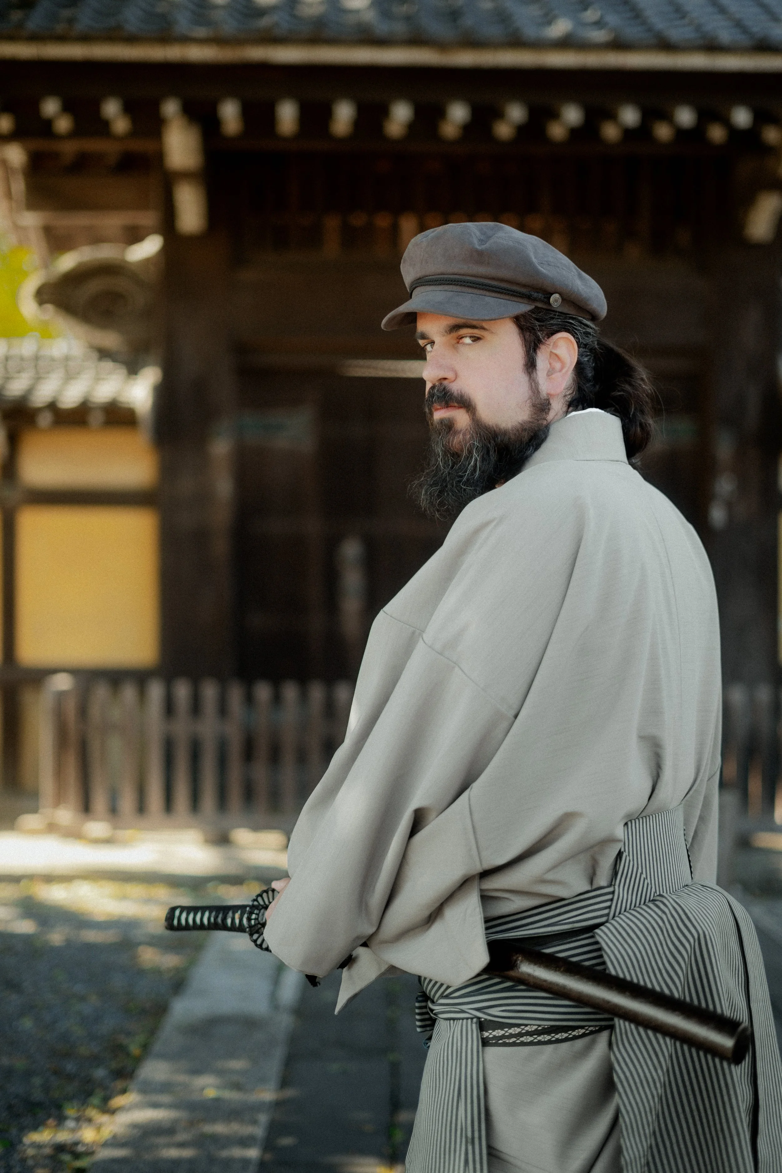 A man with long dark hair, a beard, and mustache wearing a gray cap, beige coat, and striped gray apron holding a katana sword, standing outdoors with a traditional wooden structure in the background.