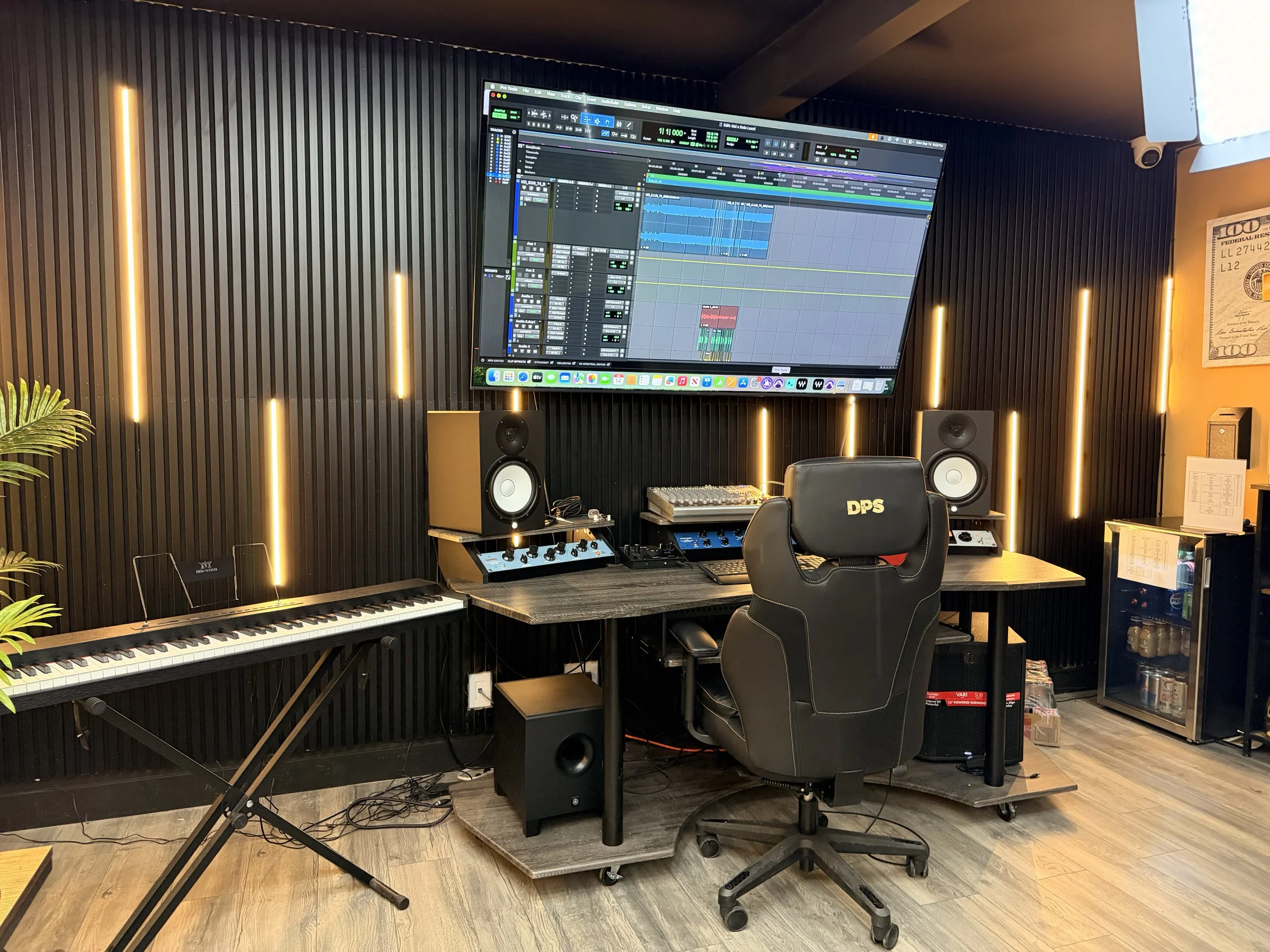Music production studio with a computer monitor displaying a digital audio workstation, a keyboard, studio speakers, a music chair, and various music equipment in a room with black acoustic wall panels and warm lighting.
