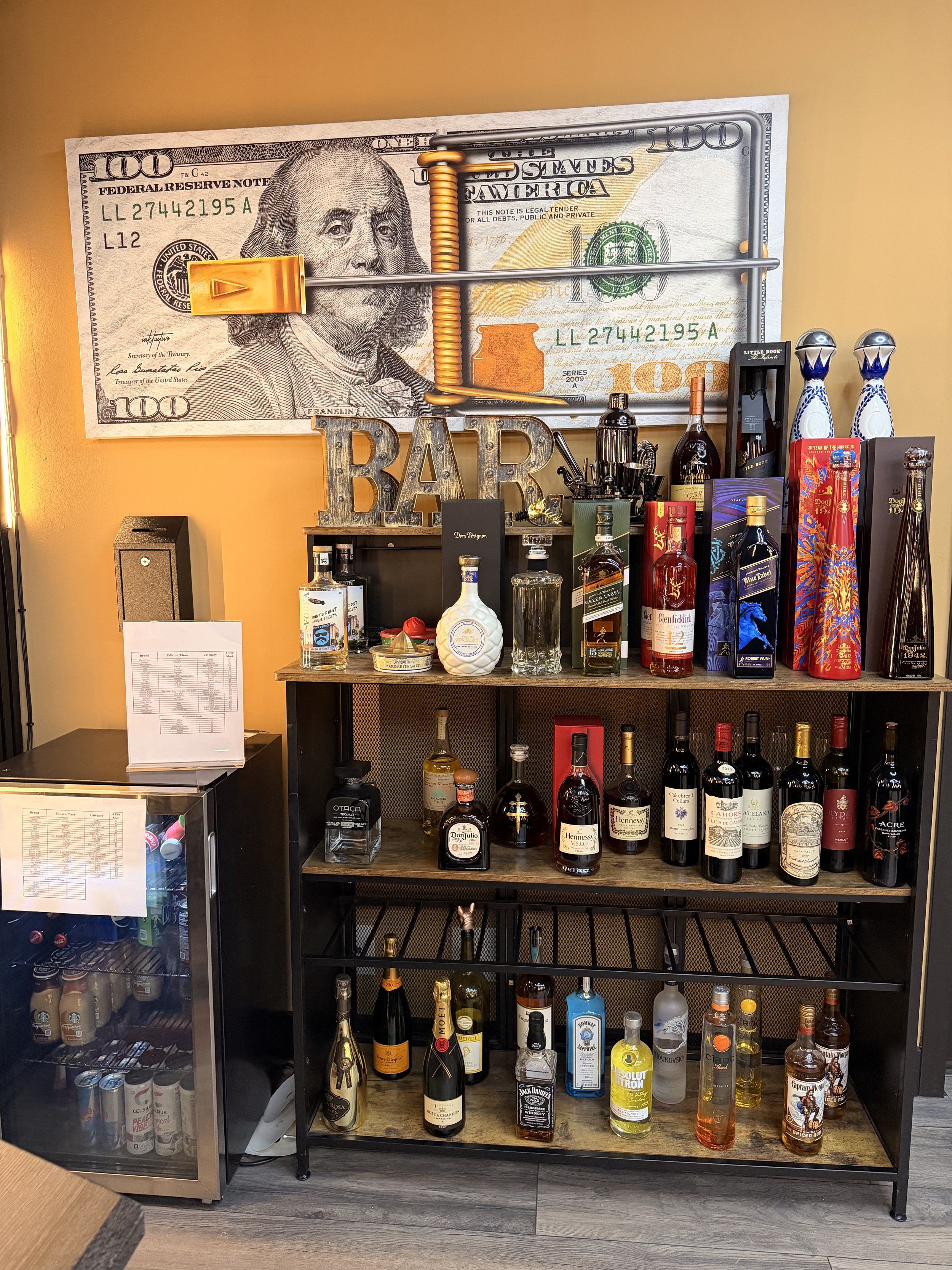 A bar shelf with various bottles of alcohol, a large dollar bill with a mouse trap on it, and a decorative sign spelling 'BAR'.