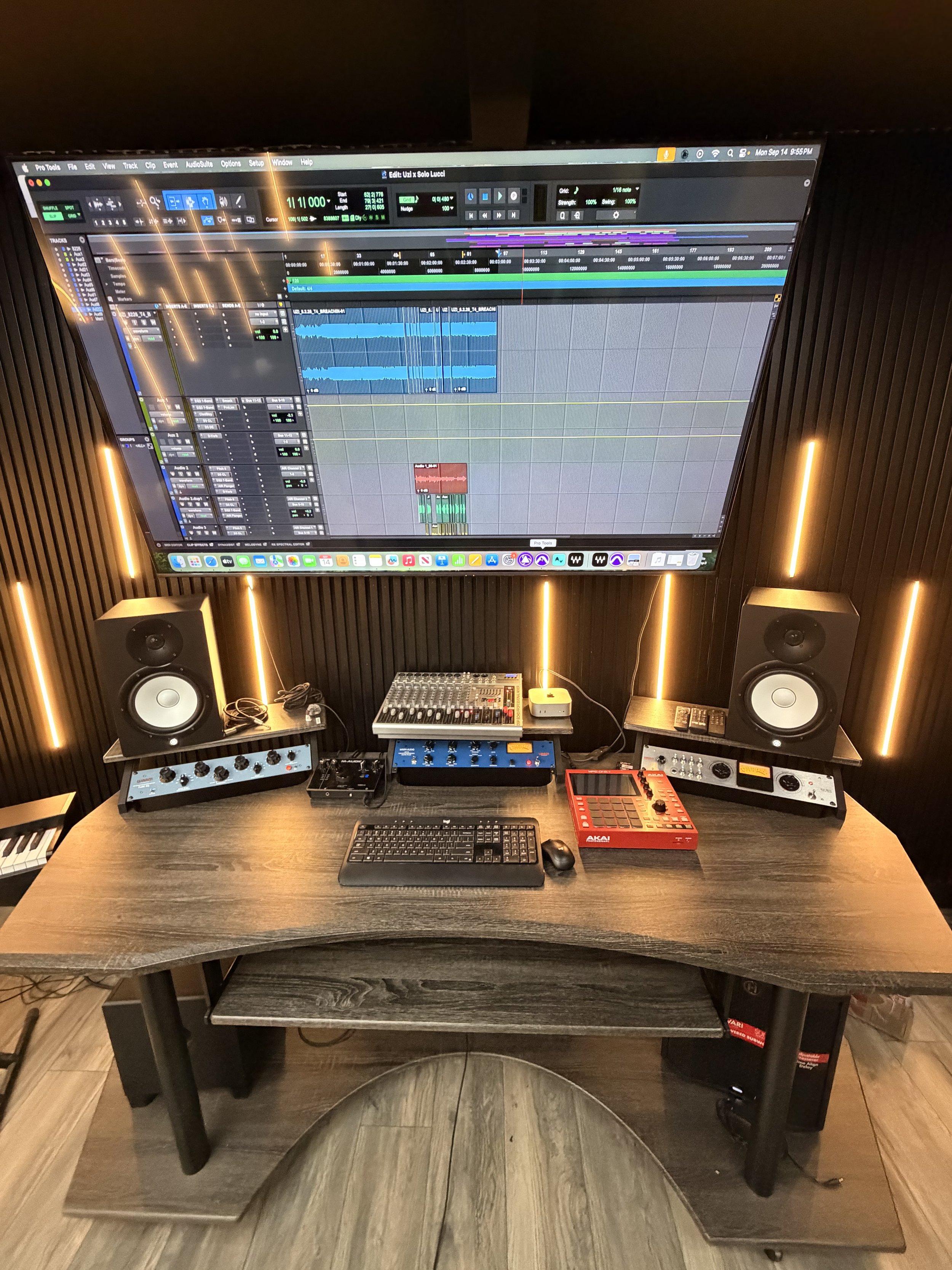 A music production setup featuring a large wall-mounted monitor displaying a digital audio workstation with multiple tracks, a desktop computer with a keyboard and mouse, a control surface, studio monitors, and a wall with black acoustic panels illuminated by vertical LED lights.