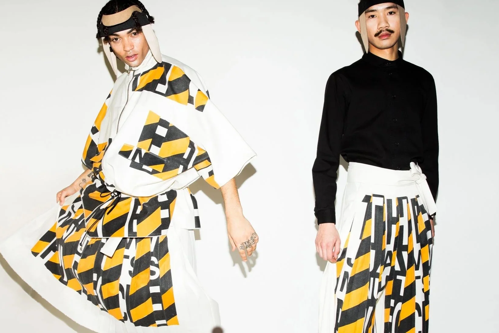 Two male models wearing the Henrik Vibskov AW16 outfits: pleated pants and an anorak with yellow, black, and white graphic prints, standing against a plain white background.