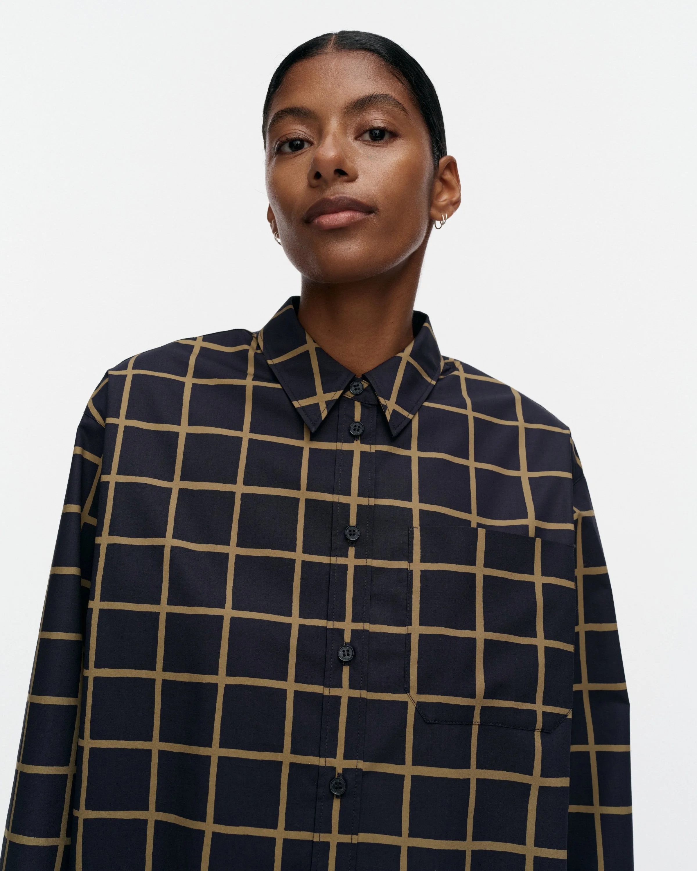 A woman with dark hair pulled back, wearing a dark button-up shirt with a beige grid pattern by Marimekko, standing against a plain white background.