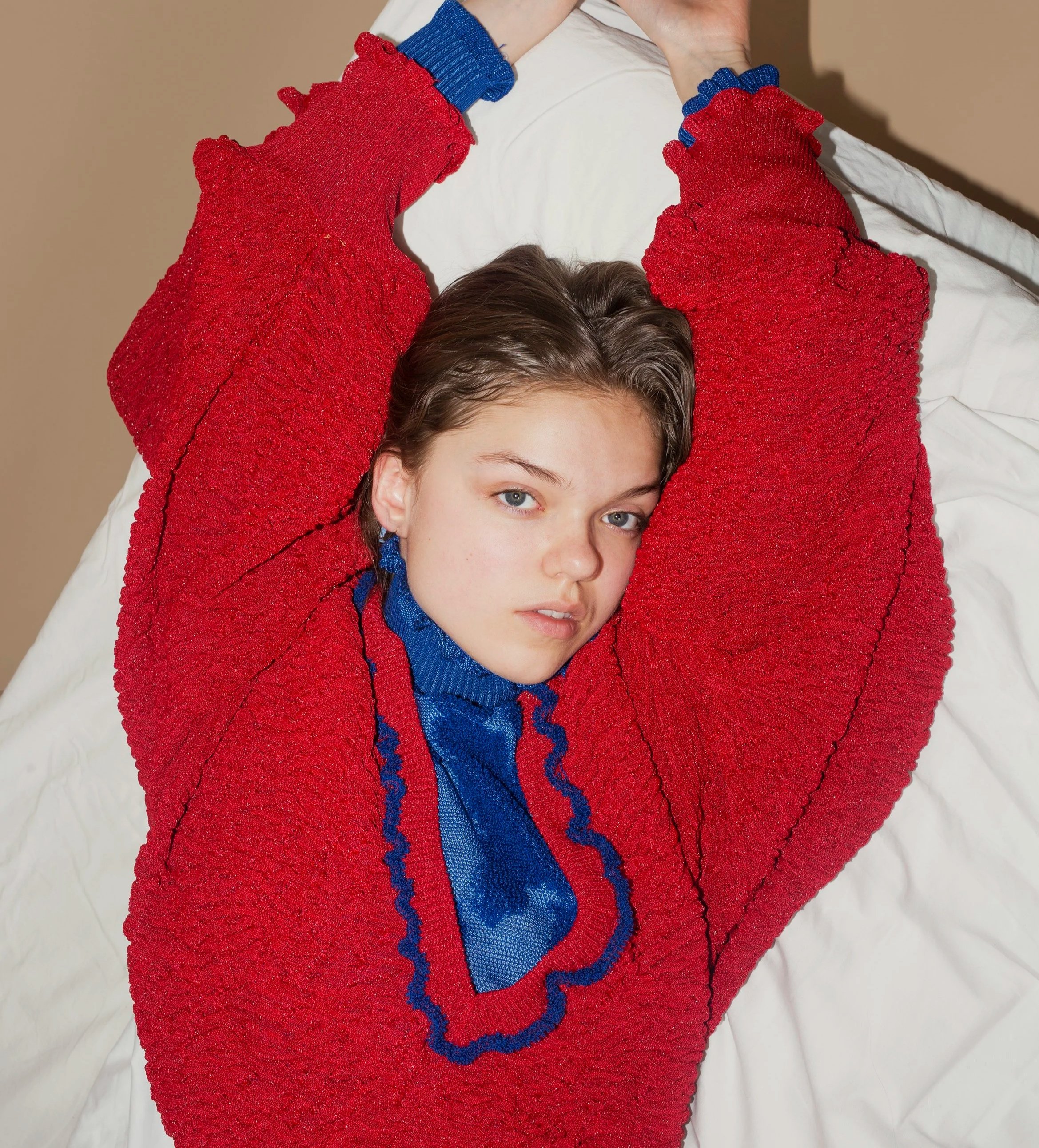 A person with short brown hair and light skin is lying on a bed with a white sheet, wearing a red hypertextured knit sweater with a v-neck collar.