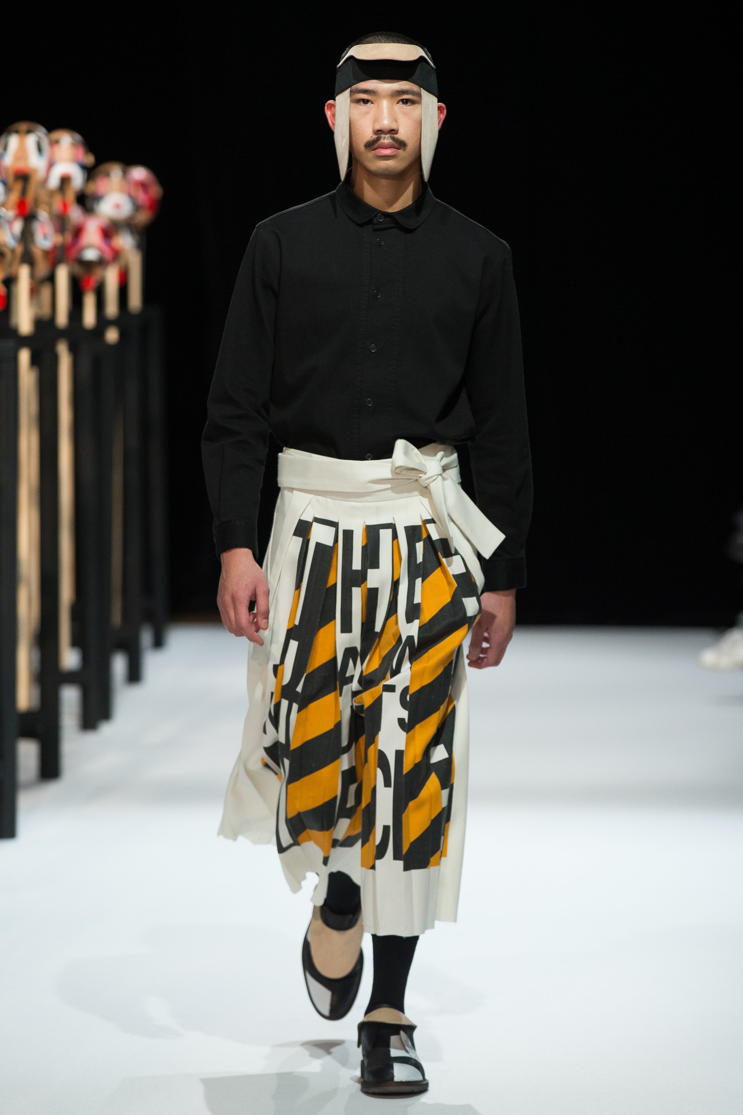 A male model walking down the Paris Men's Fashion Week runway for the Henrik Vibskov AW16 show, wearing a black button-up shirt, white pants with large text print in black, yellow, and gray.