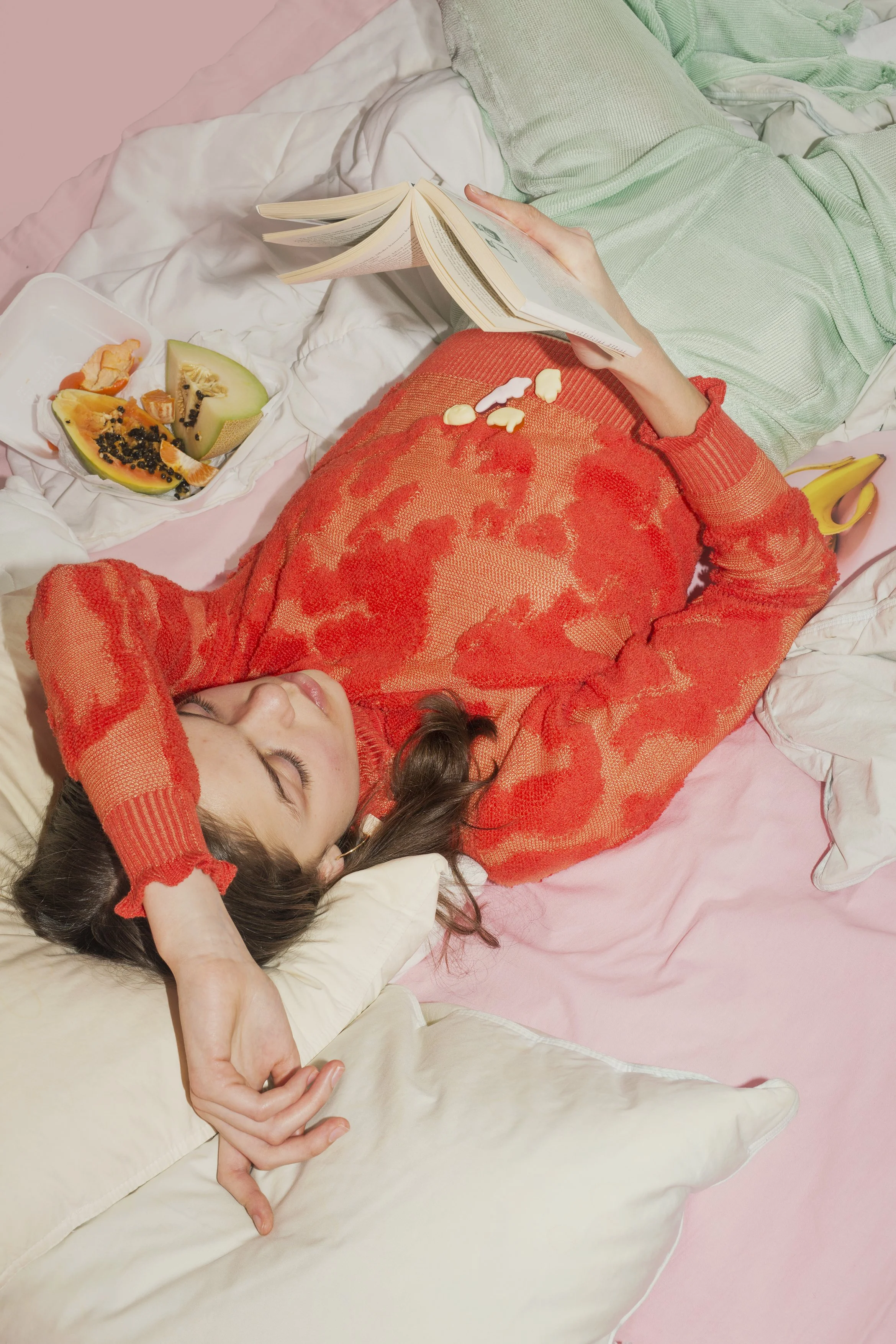 A woman lying on a bed with pink sheets, wearing an orange hypertexturized knit sweater and glossy mint-color knit pants, reading a book, with a bowl of cut fruit including papaya, melon, and grapes.
