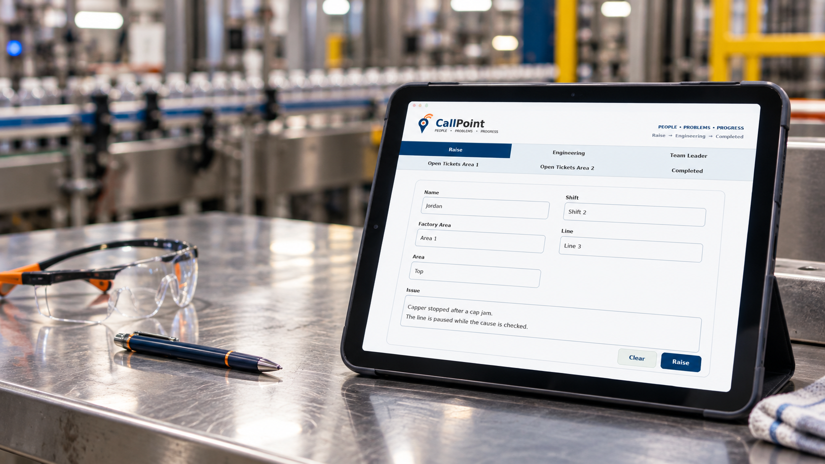 Tablet on a metallic table showing a factory support ticket system, with a pair of safety glasses and a pen nearby, in an industrial setting.