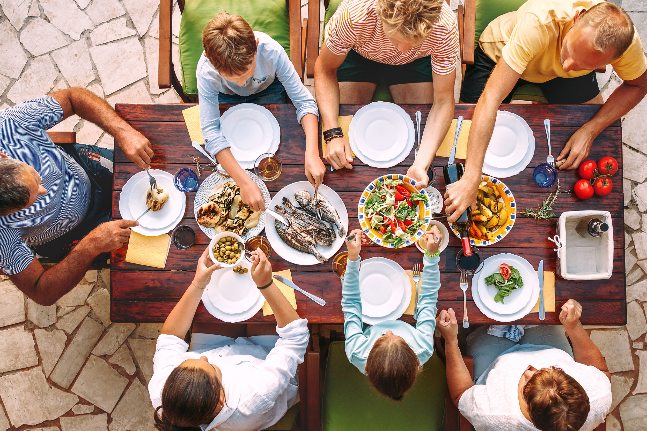 An overhead view of a family gathering around a wooden dining table outdoors. The table is set with white plates, cutlery, and glasses. A variety of foods, including a salad, grilled fish, roasted vegetables, bread, olives, and tomatoes, are on the table. The people are reaching for food and serving themselves, with some wearing casual clothing. The background shows a stone patio surface.