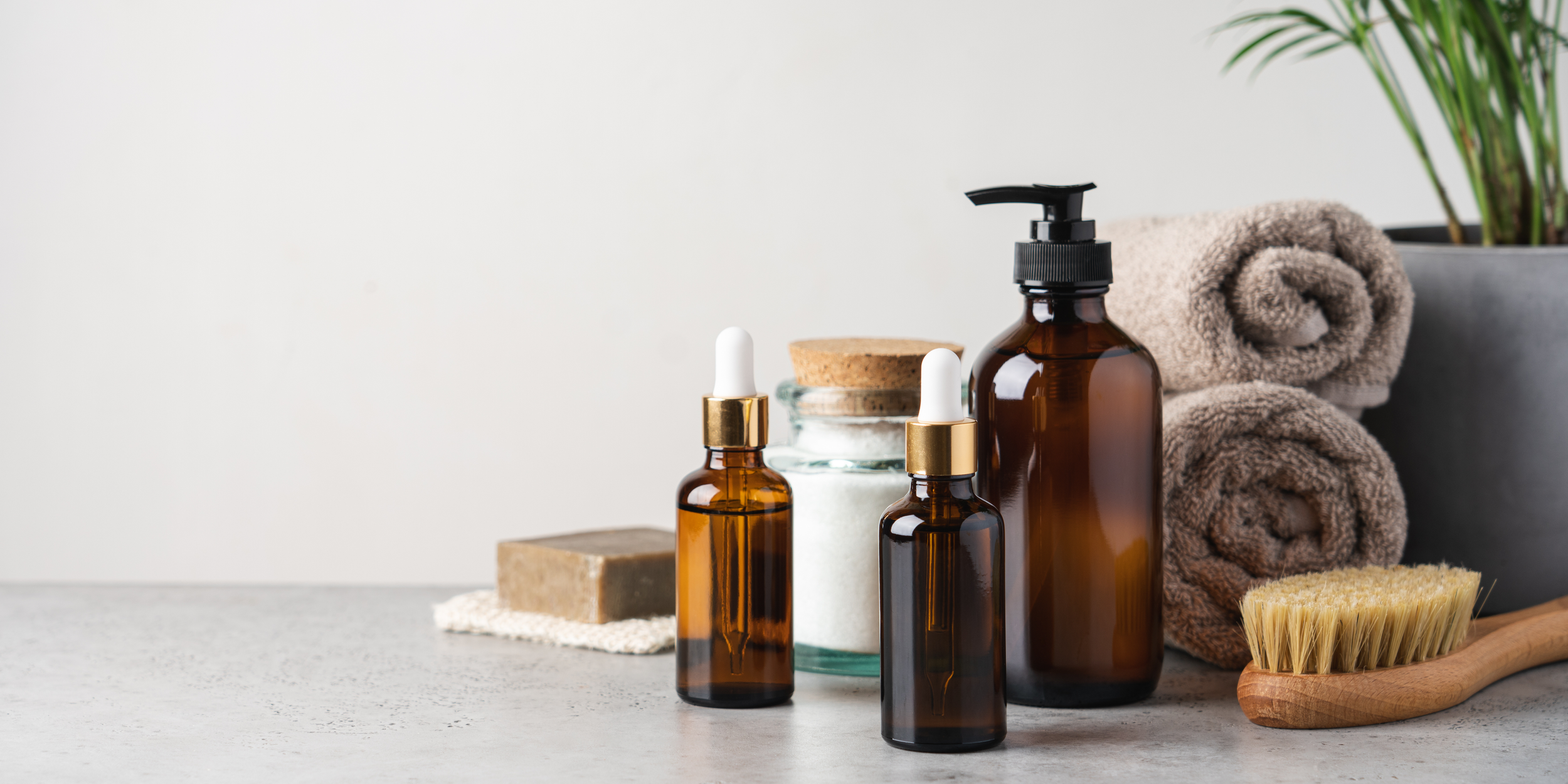 Home and wellness products, including amber glass bottles with droppers, a pump bottle, rolled towels, a potted plant, a brush, and soap on a light surface.