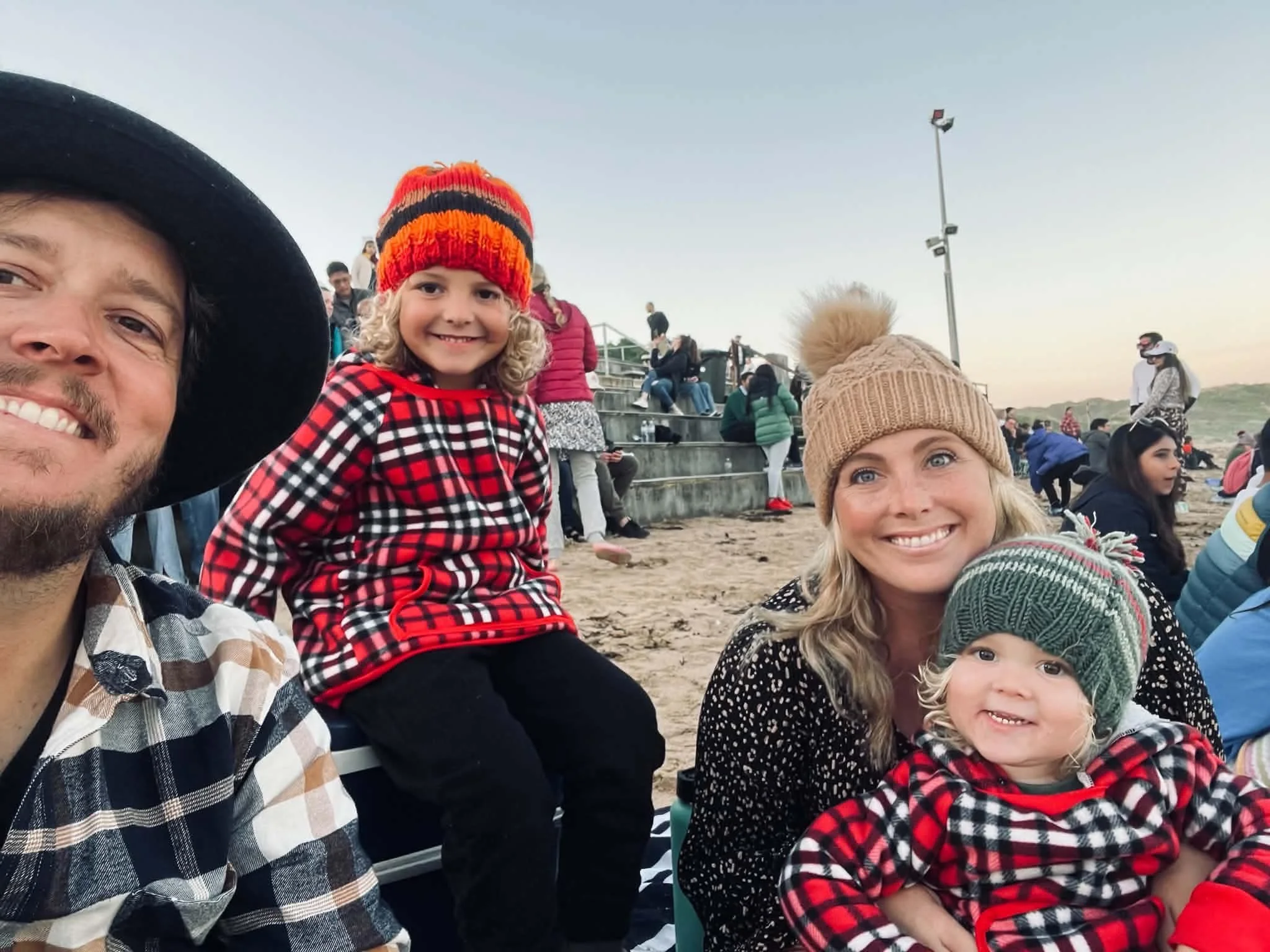 The Loats family, including children and adults, is sitting outdoors on a sandy area with bleachers and other people in the background, during what appears to be an event or gathering at sunset.