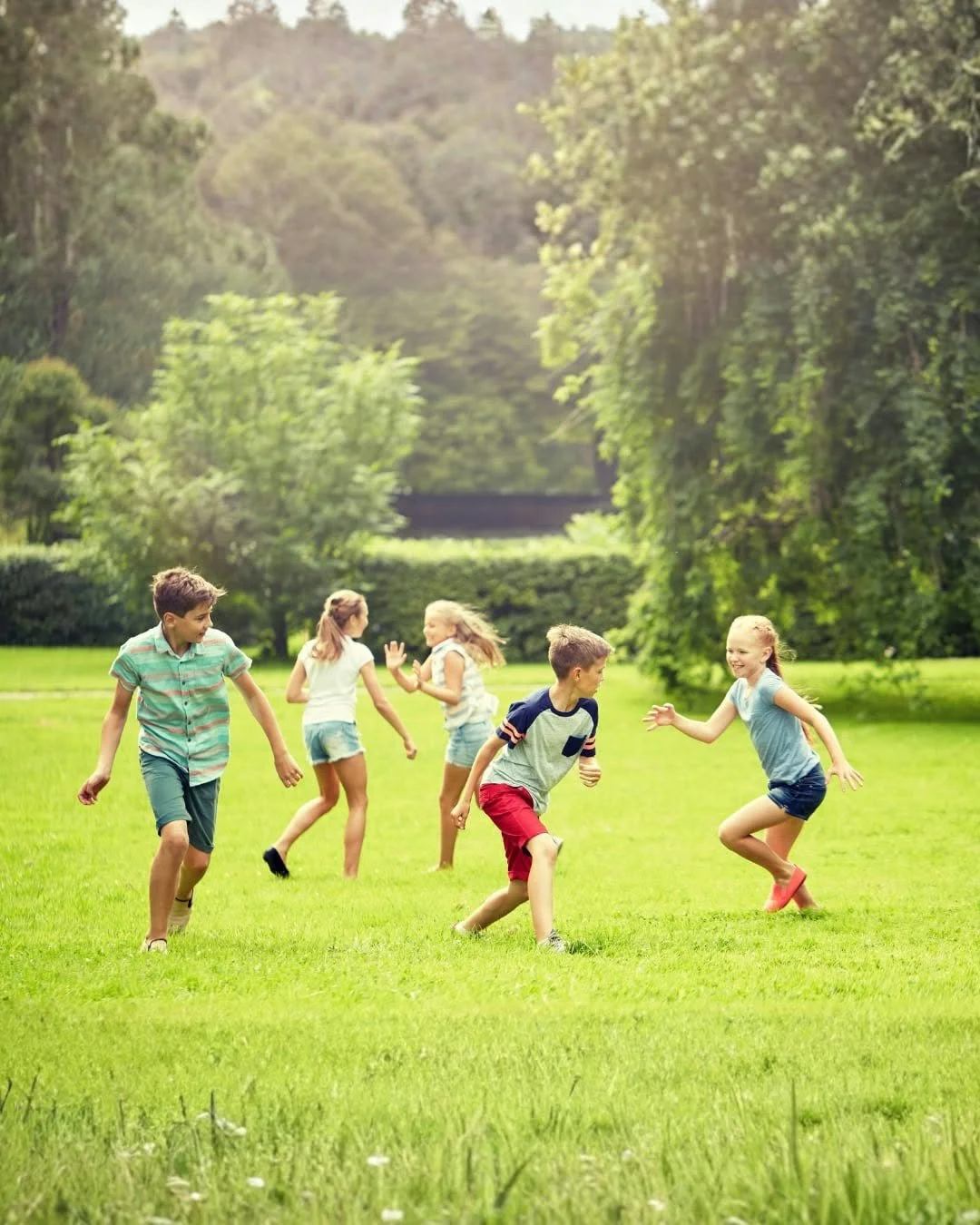 Children playing and running in a grassy park with trees and a bridge in the background during daytime.