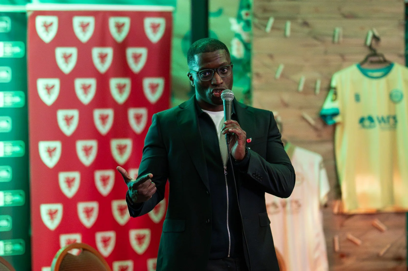 Daunte Crawford in a black suit with glasses holding a microphone and speaking, standing in front of a red banner with shield-shaped logos and a yellow sports jersey hanging on the wooden wall.